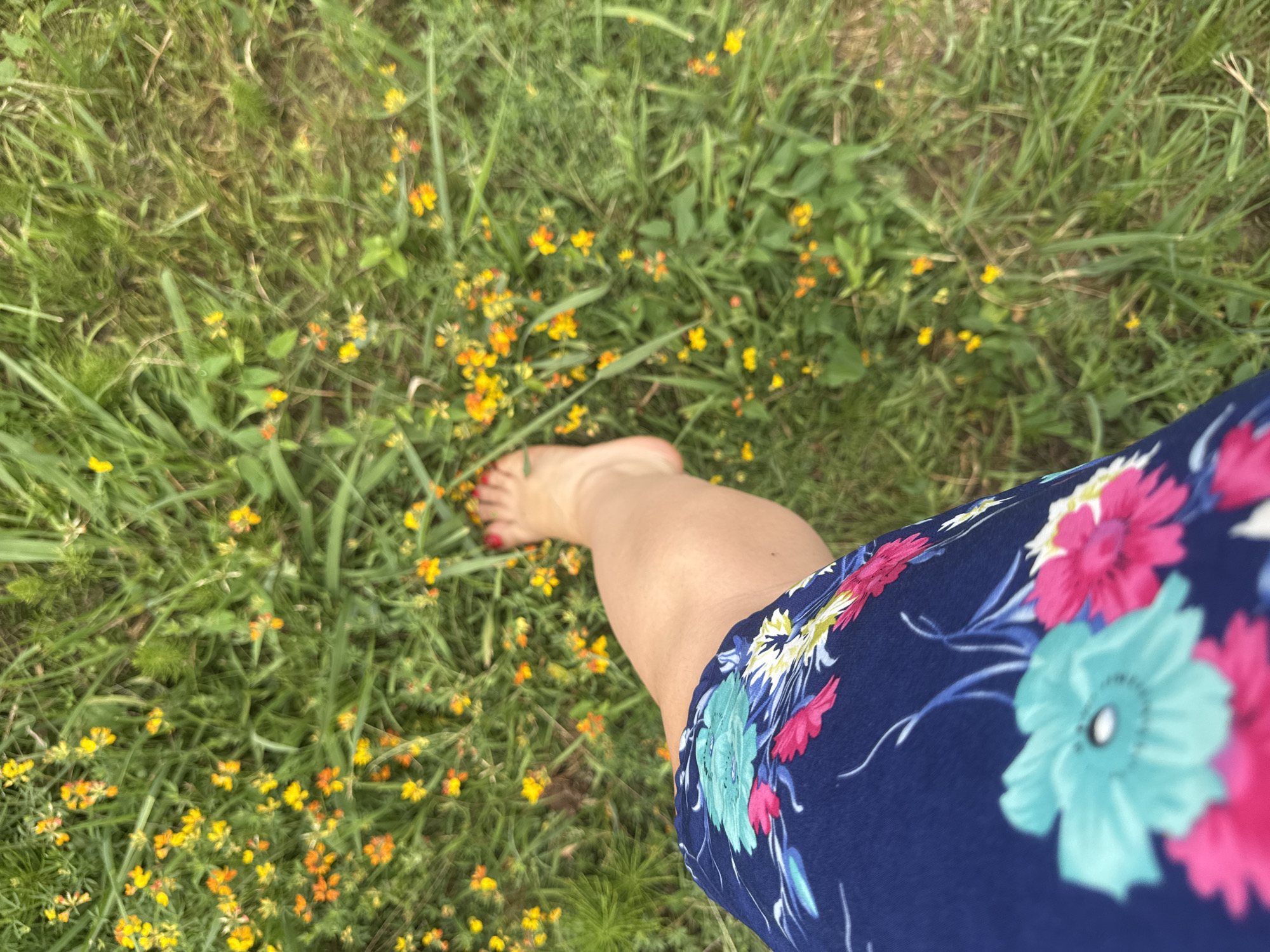 The image shows a close-up, outdoor shot of a person's bare foot and lower leg, wearing a dark blue dress with a floral pattern. The foot is on green grass with scattered small yellow and orange flowers. The setting is likely a park or garden. There is no visible indication of the location being the city of Sochi, Russia.
