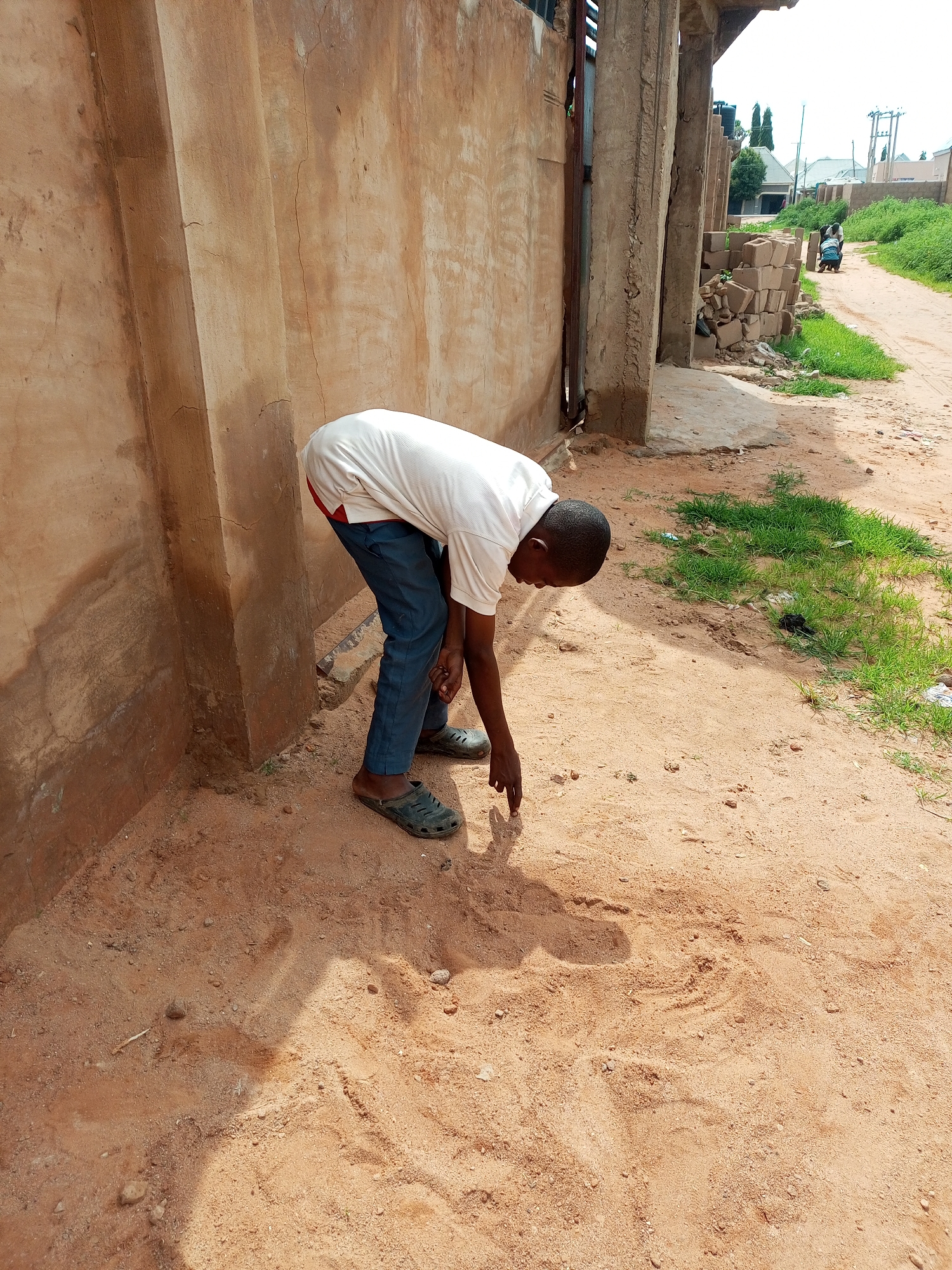 The image shows a young Black male, wearing a white t-shirt with red trim at the collar, dark blue trousers, and dark-colored sandals, bending over and pointing at the sandy ground. He is positioned in the foreground, to the left of the frame, next to a rough, tan-colored wall. The ground is covered in reddish-brown sand with scattered small stones and some dry grass. To the right of the sandy area, there is a dirt road with some green grass growing along its edges. In the background, there are buildings, vegetation, and utility poles, suggesting a semi-urban or rural environment. The overall scene depicts a moment of observation or interaction with the ground.