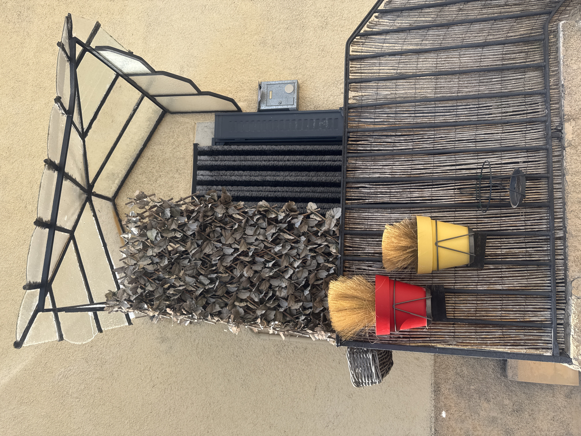 This image shows an outdoor scene in Saint-Claude, France, likely a residential exterior. The focus is on a collection of objects and architectural elements attached to a textured, light beige wall.
Prominently featured is a unique canopy structure on the left, constructed with a metal frame and panels of translucent material, possibly glass or plastic, arranged in a leaf-like or fan-like pattern. Below this canopy, a dense cluster of dried, brown leaves is visible, forming a textured mat or covering.
To the right of the leaves, a dark grey doormat or grate is positioned, with horizontal slats. Above the doormat, a small, grey metal box, possibly a fuse box or electrical panel, is mounted on the wall.
Further to the right, a larger structure made of horizontally aligned, natural-colored reeds or bamboo, framed by dark metal, forms a screen or shade. Attached to this reed structure are two decorative items that resemble brooms or brushes. One has a bright yellow container, and the other has a red container. Both have light brown bristles extending outwards. A small, woven basket hangs below the red container. There is also a black wire loop and a dark, shallow bowl-like object mounted on the reed screen.
The lighting suggests it is daytime, with natural light illuminating the scene. The wall texture and the reed screen provide cues about the location possibly being in a warmer climate or a region with specific architectural styles. No people are visible, and the overall mood is quiet and still. There is no visible text in the image.