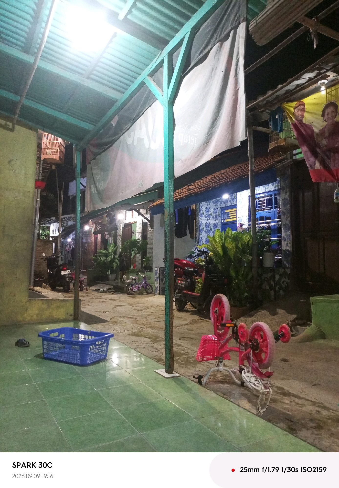 An exterior shot captures a dimly lit alleyway in Tegal, Indonesia, under a corrugated tin roof supported by teal metal beams. The scene is cluttered with various objects, suggesting a residential or workshop area. In the foreground, a bright blue plastic basket rests on a green tiled floor. To the right, a pink child's tricycle with red wheels is overturned, its basket facing upwards. A dark motorcycle is parked on the left, and another red motorcycle is visible further back. The background reveals multiple doorways and windows of houses, some illuminated by interior lights. A large white banner with black lettering hangs across the top of the alley. Laundry is seen hanging on a clothesline in the middle ground. A birdcage is perched on a wooden structure on the upper left side of the image. The overall impression is one of a lived-in, functional space in an urban setting.