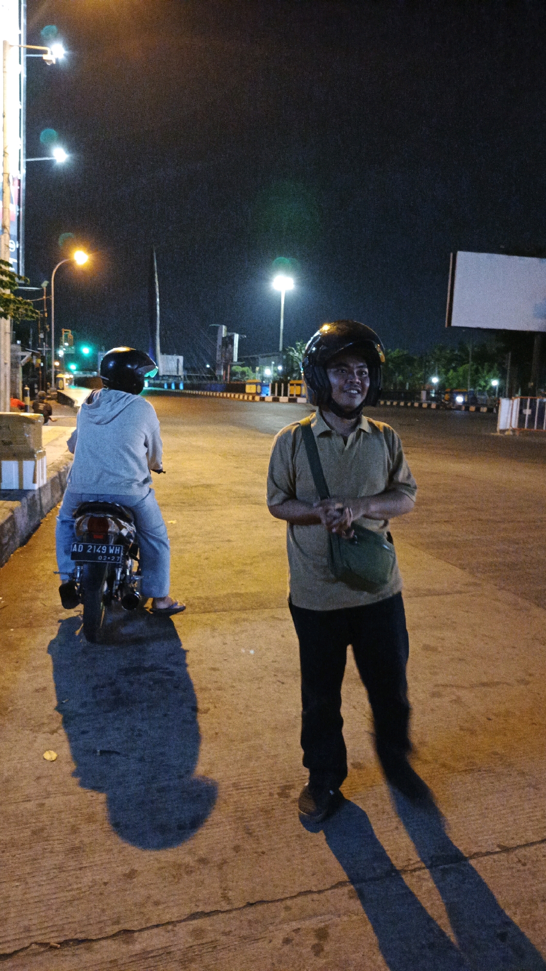 Two individuals are standing on a paved street at night in the city of Surakarta, Indonesia. The person on the left is seated on a motorcycle, facing away from the camera, and is wearing a light gray hooded sweatshirt, light blue pants, and a dark helmet. The motorcycle has a license plate that reads "AD 2149 WH". The person on the right is standing and facing the camera, wearing a light brown collared shirt, dark pants, a dark helmet, and a green satchel across their chest. They appear to be speaking or posing for the camera. The background shows streetlights, traffic signals, and buildings, suggesting an urban environment. The lighting is artificial and casts long shadows on the ground.