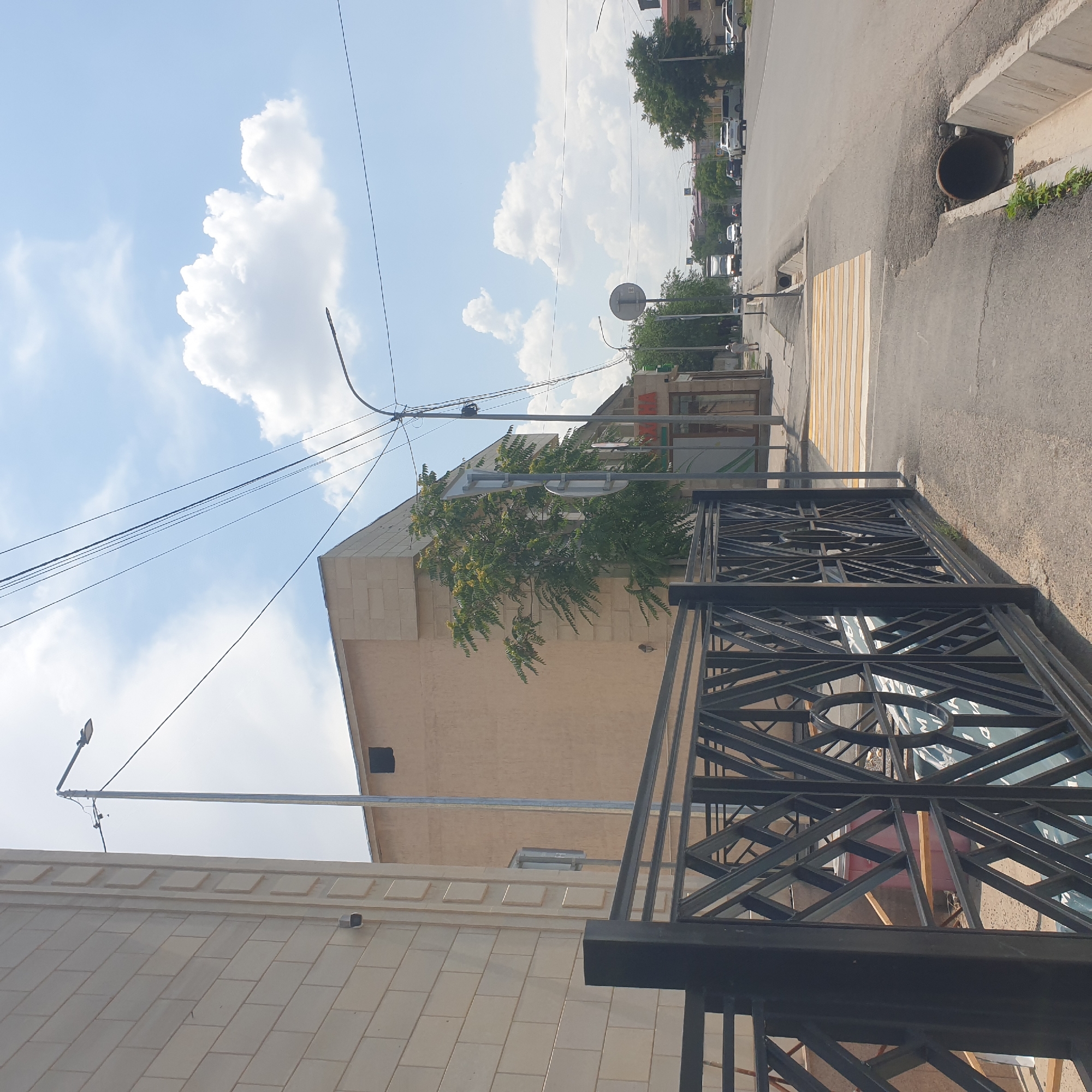 The image depicts a street scene in Shymkent, Kazakhstan, on a bright, partly cloudy day. In the foreground, a black metal decorative fence runs along the left side of the frame, with a concrete sidewalk adjacent to it. A pedestrian crosswalk with yellow and white stripes is visible across the street, leading to the right. A concrete drainage ditch with a large pipe entering it is present on the right side of the road.
On the left, a building with light-colored brick-like cladding is visible, featuring a security camera mounted on its facade. Next to it, a tan-colored building with a dark square opening is present. A tall metal pole with a street light is situated between these buildings, with power lines extending across the sky. Further down the street, a tree with green foliage stands near another building.
The street itself is paved and appears to be a moderately busy road with several cars and a truck visible in the distance. Traffic lights and street lamps are present along the route. In the background, more buildings and trees are scattered throughout the urban landscape. Two individuals are visible: one person walking on the sidewalk in the distance on the right, and another person standing near a building on the left side of the street. The sky is a clear blue with scattered white cumulus clouds.