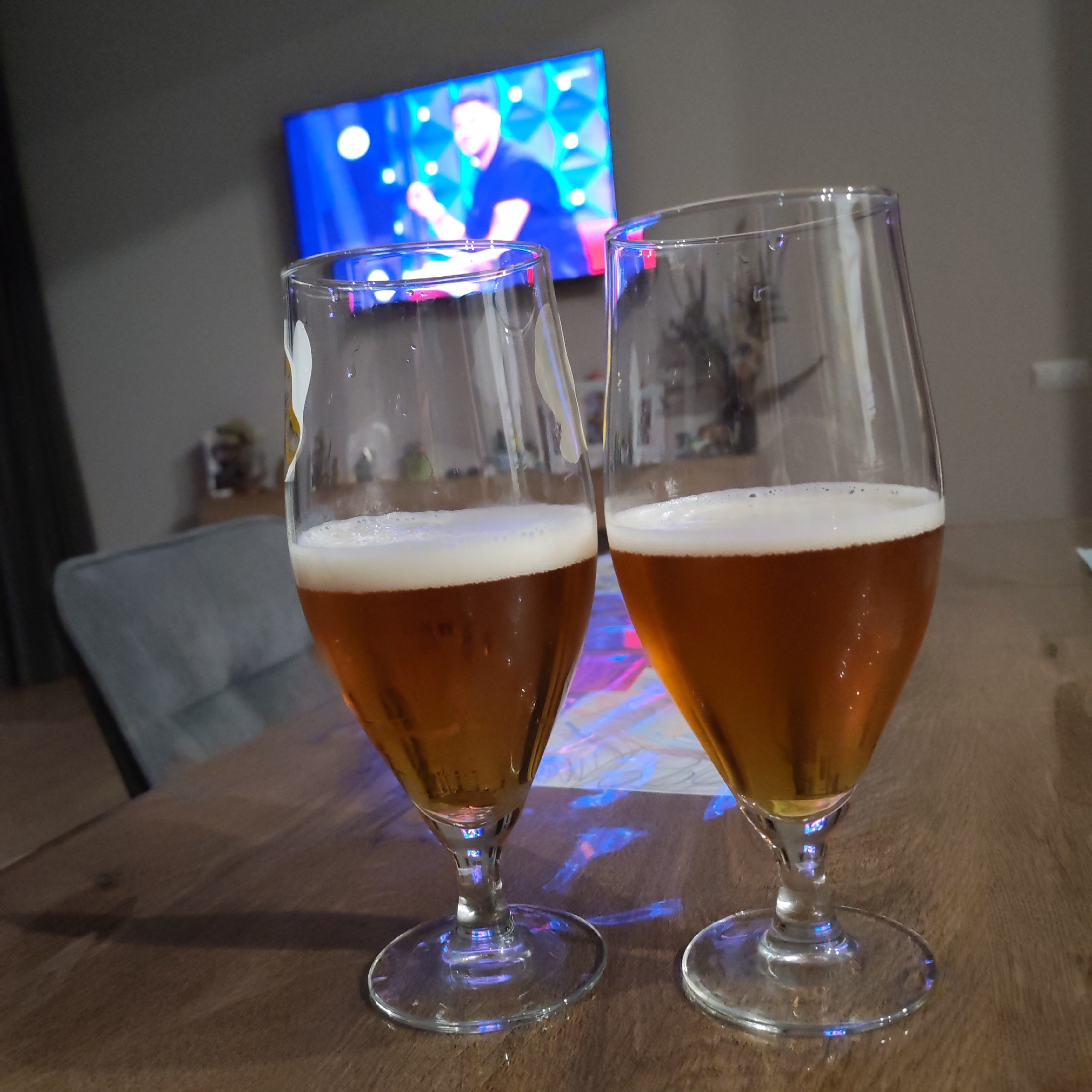Two glasses of amber-colored liquid with foamy tops sit on a wooden table. In the background, a television screen displays a man in a dark shirt, with bright blue and white lights behind him. The setting appears to be indoors, possibly a living room or dining area, with a grey armchair visible to the left and a neutral-colored wall behind. The lighting suggests it is evening. There is no indication that this scene is located in Балаклавский округ, Ukraine.