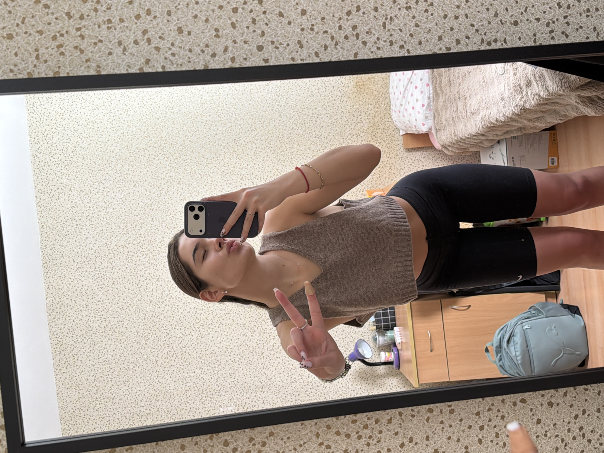 A young woman takes a selfie in a mirror. The mirror reflects a room, which includes a bed, a desk with drawers, and a backpack. The woman is wearing a brown top and black shorts. She is holding a phone in one hand and making a peace sign with the other. The location is Sofia, Bulgaria.