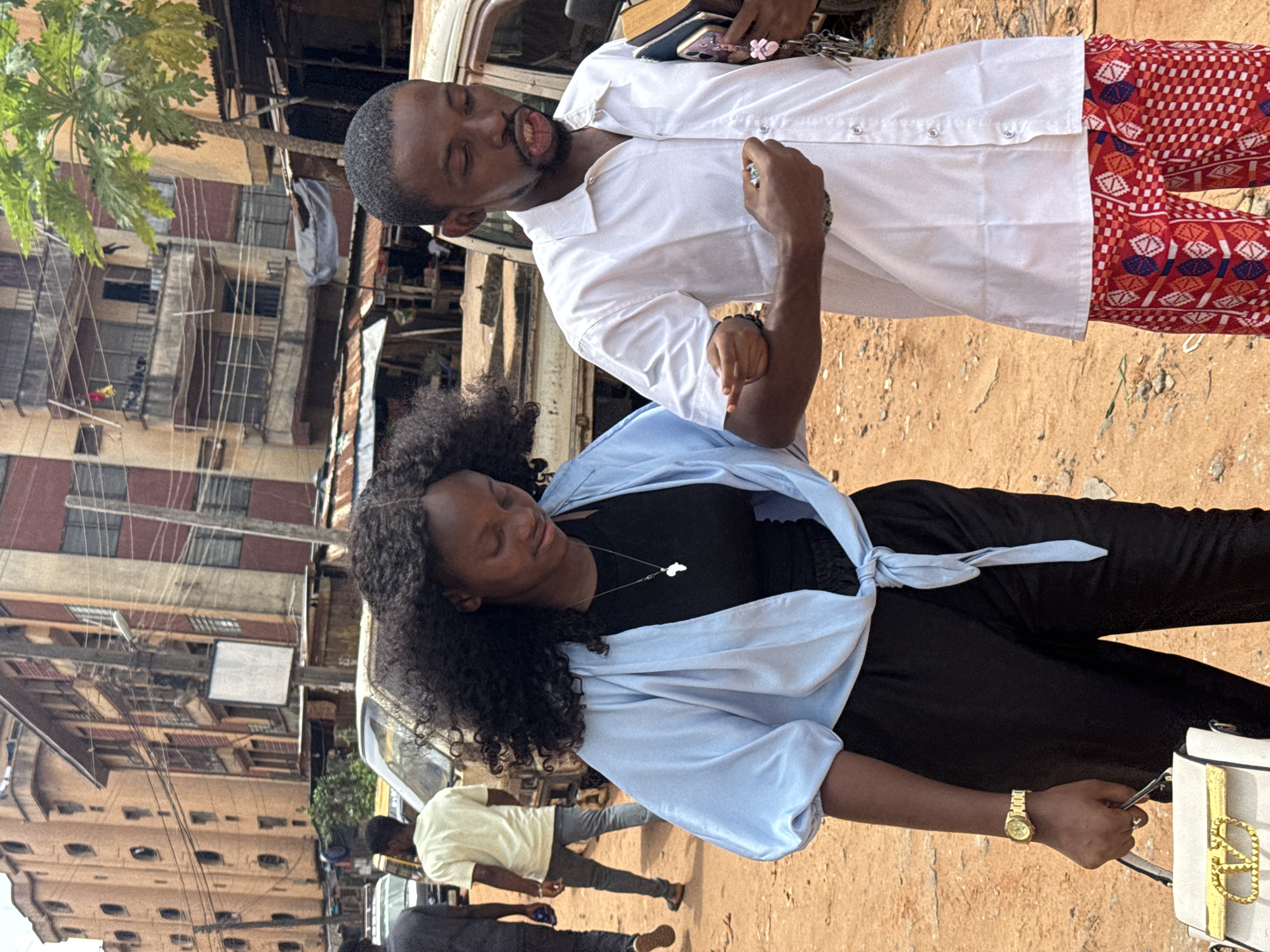 The image shows a man and a woman standing in an urban environment in Onitsha, Nigeria. The man is wearing a white short-sleeved shirt, and red pants with geometric patterns. He is holding keys and a mobile phone. The woman is wearing a black top, black pants, and a light blue shirt. She is holding a white purse and wearing a gold watch. Buildings are visible in the background, along with a partially visible vehicle. Other people are walking nearby. The ground appears to be dirt.