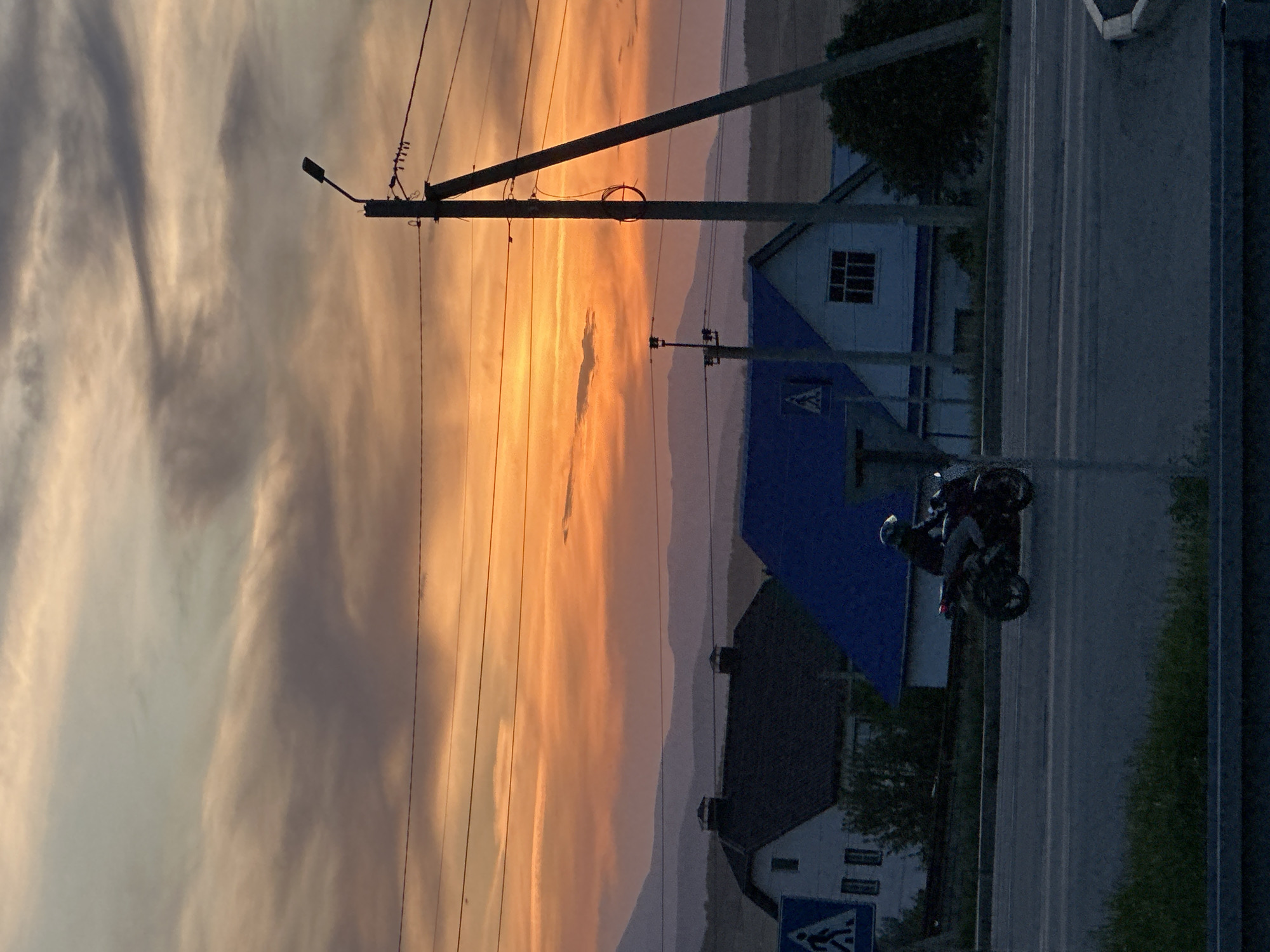 The image shows a street scene at dusk. A motorcycle with two riders is on the road, moving away from the viewer. Buildings are on the roadside, with a house having a distinctive blue roof. A telephone pole stands near the road. The background features a sky with shades of orange and gray, likely indicating sunset or sunrise. The location is an urban environment.
