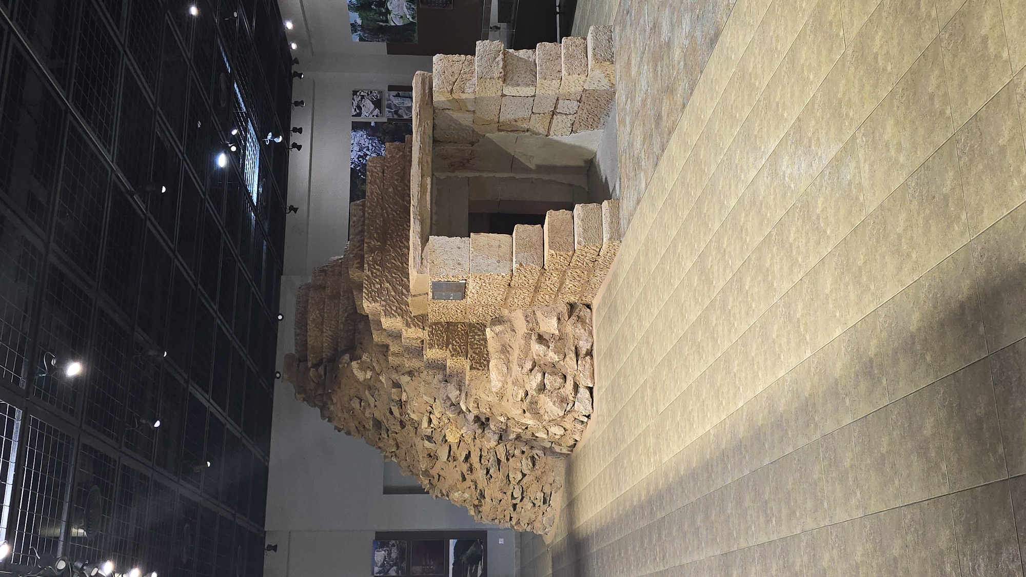 The image depicts the interior of a museum exhibit, showcasing a reconstruction of an ancient structure resembling a stepped pyramid with an entrance. The structure is made of rough-hewn stone and earth-like materials, exhibiting a weathered and ancient appearance. Steps lead up the left side of the structure, while a doorway with a stone lintel provides access to the interior on the right. The floor is tiled with large, rectangular light brown tiles, arranged in a grid pattern, with shadows cast across it. The ceiling features a dark grid structure with spotlights illuminating the exhibit. In the background, there are informational displays with images on the walls, and other parts of the museum are visible, including a glass display case. The overall environment suggests a historical or archaeological exhibition. The provided context indicates this exhibit is located in the city of Konstantinovo, Bulgaria. There are no people visible in the image.
