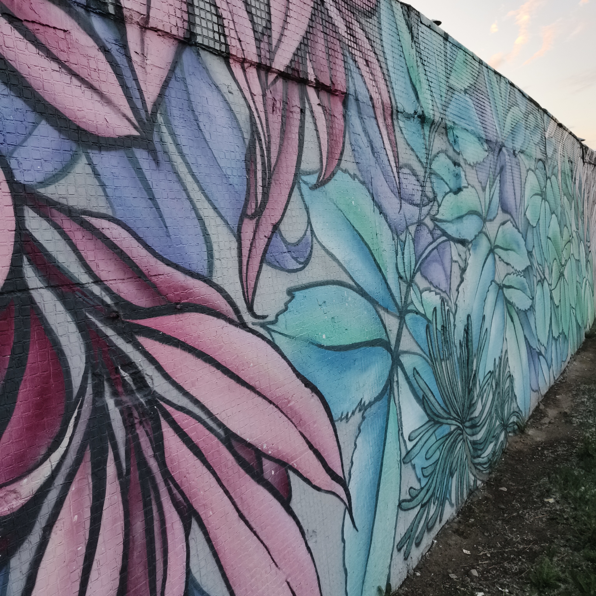 The image shows a graffiti mural on a brick wall, depicting large, stylized leaves in shades of pink, purple, blue, and green. The mural extends along the length of the wall, with the colors transitioning from pink and purple on the left to blues and greens on the right. The leaves are outlined in black. The wall appears to be outdoors, possibly an exterior wall of a building or a sound barrier, with a dirt and grass embankment visible at the base of the wall. The sky is visible in the upper right corner, with some clouds. The location is identified as Chaikovsky, Russia. No people are present in the image.