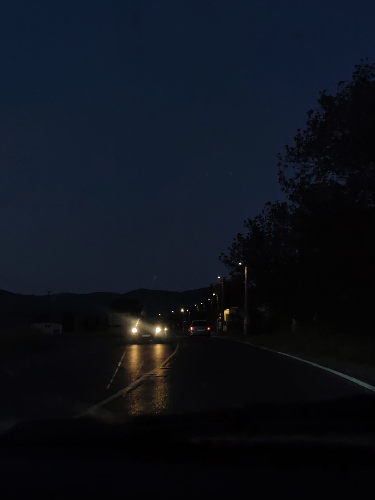 A nighttime photograph depicting a road with vehicle traffic. Two cars with headlights illuminated are visible in the distance. Another car with red taillights is also visible farther down the road. The road appears wet, reflecting light from the vehicles and streetlights. Trees line the right side of the road, obscuring objects behind them. Hills are visible in the distance. Streetlights illuminate the road. The sky is dark. The location appears to be a rural or suburban area.