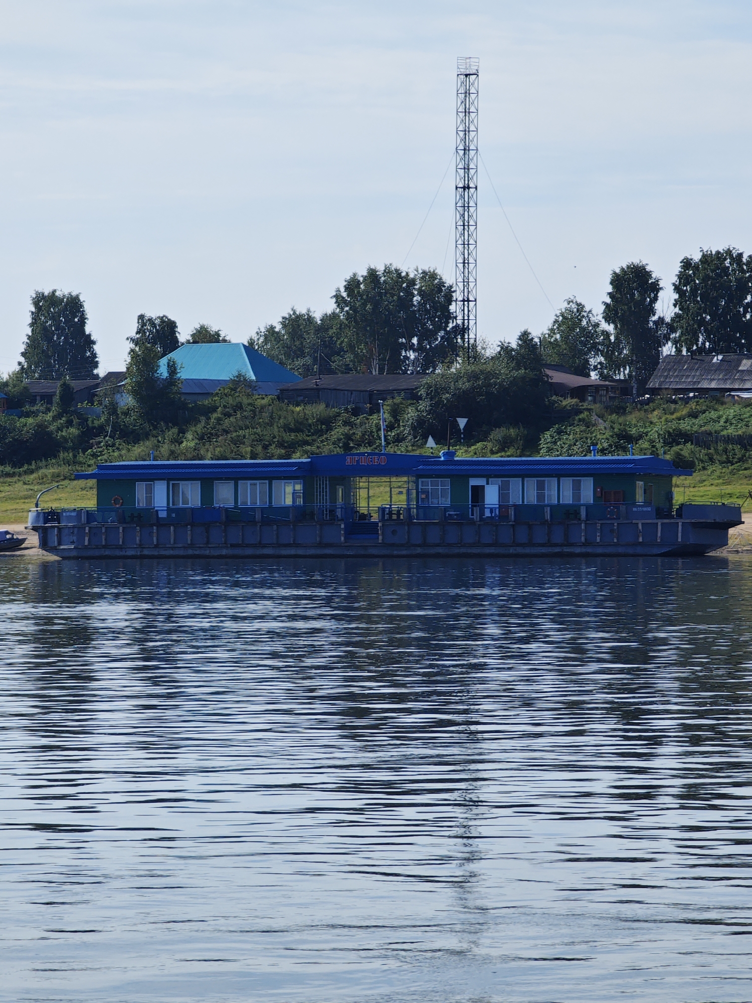 This image depicts a waterfront scene in the city of Yarstevo, Russia. In the foreground, a calm body of water reflects the sky and the objects on the shore. A long, flat-bottomed boat with a blue roof and green walls is moored along the bank. The boat has several windows and appears to be a passenger vessel or a floating structure. Behind the boat, a line of trees and some buildings are visible, suggesting a semi-urban or rural setting. A tall, lattice-like communication tower stands prominently against the sky. The overall environment is bright and suggests a daytime scene under a clear or partly cloudy sky.