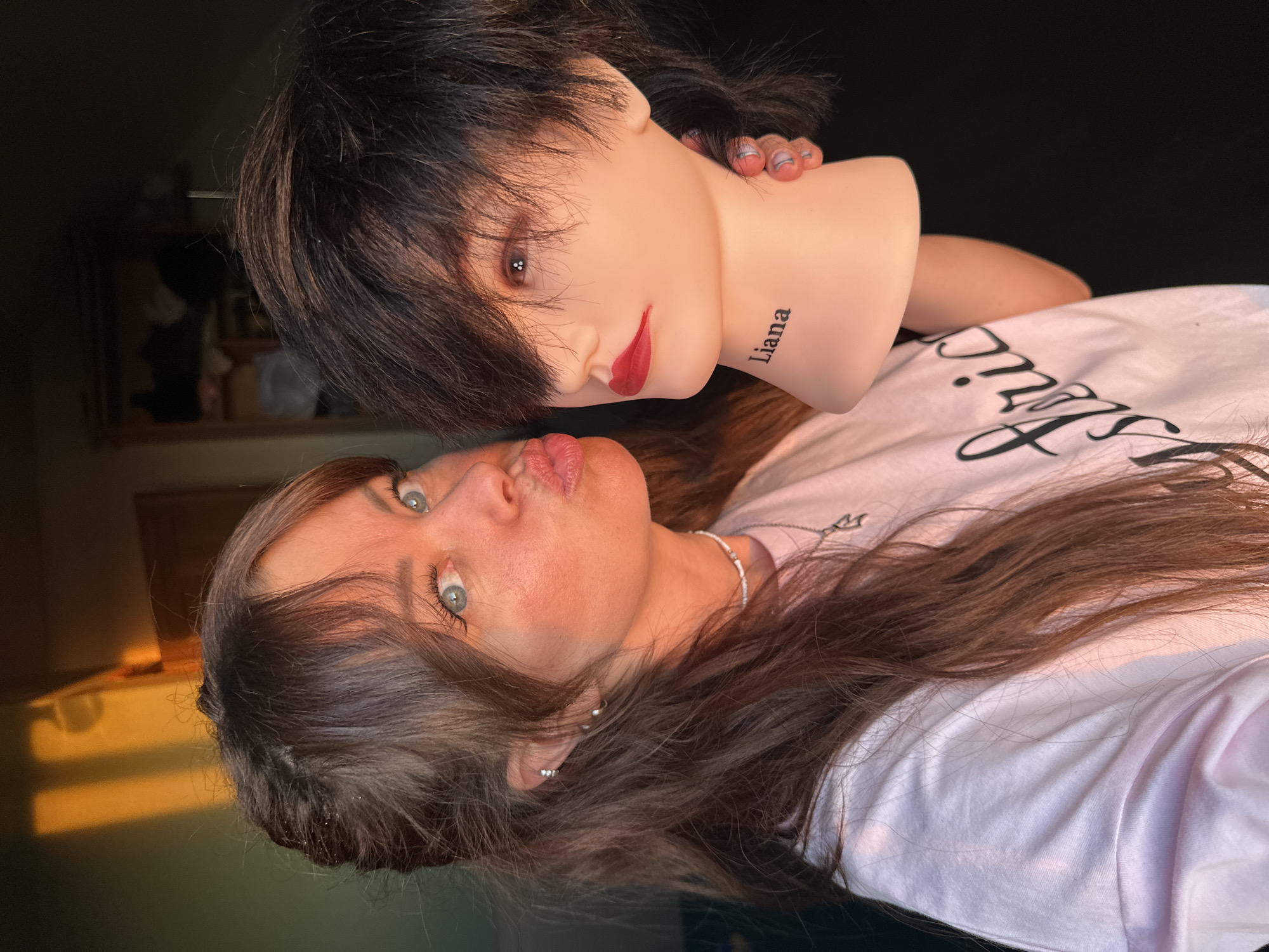 A light-skinned woman with long, wavy brown hair and blue eyes is posing with a mannequin head. The woman is wearing a white t-shirt and silver earrings, and has her lips pursed as if giving a kiss. She also wears a white bead necklace with a silver charm. The mannequin head has short, black hair with bangs, red lipstick, and the name "Liana" tattooed on its neck. The woman is holding the mannequin head. The background is a dark, indistinct room with a lighter area behind her. The location is not discernible as Chaikovsky, Russia, based on the visual details in the image.