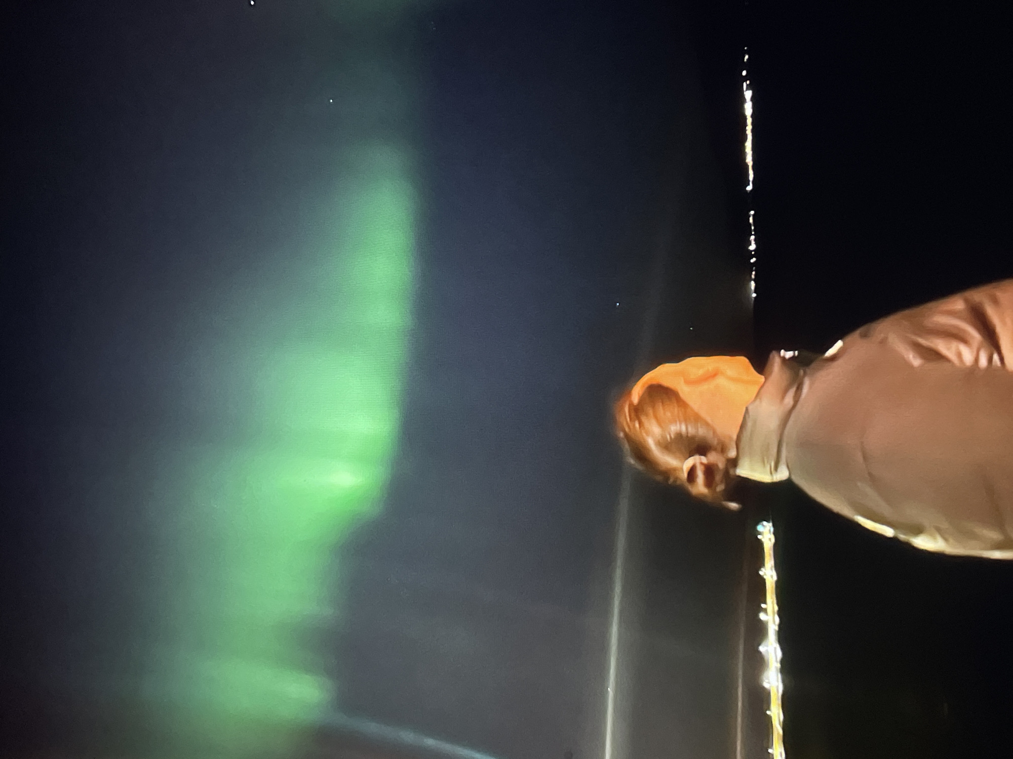 The image shows a person, with reddish-blonde hair, wearing a tan coat. The person is seen against a backdrop of the aurora borealis, which appears as a green, hazy band in the sky. A string of lights is visible near the person. The location appears to be a dark area, with the aurora and string lights providing the only light sources. The location in the city is unknown, unknown.