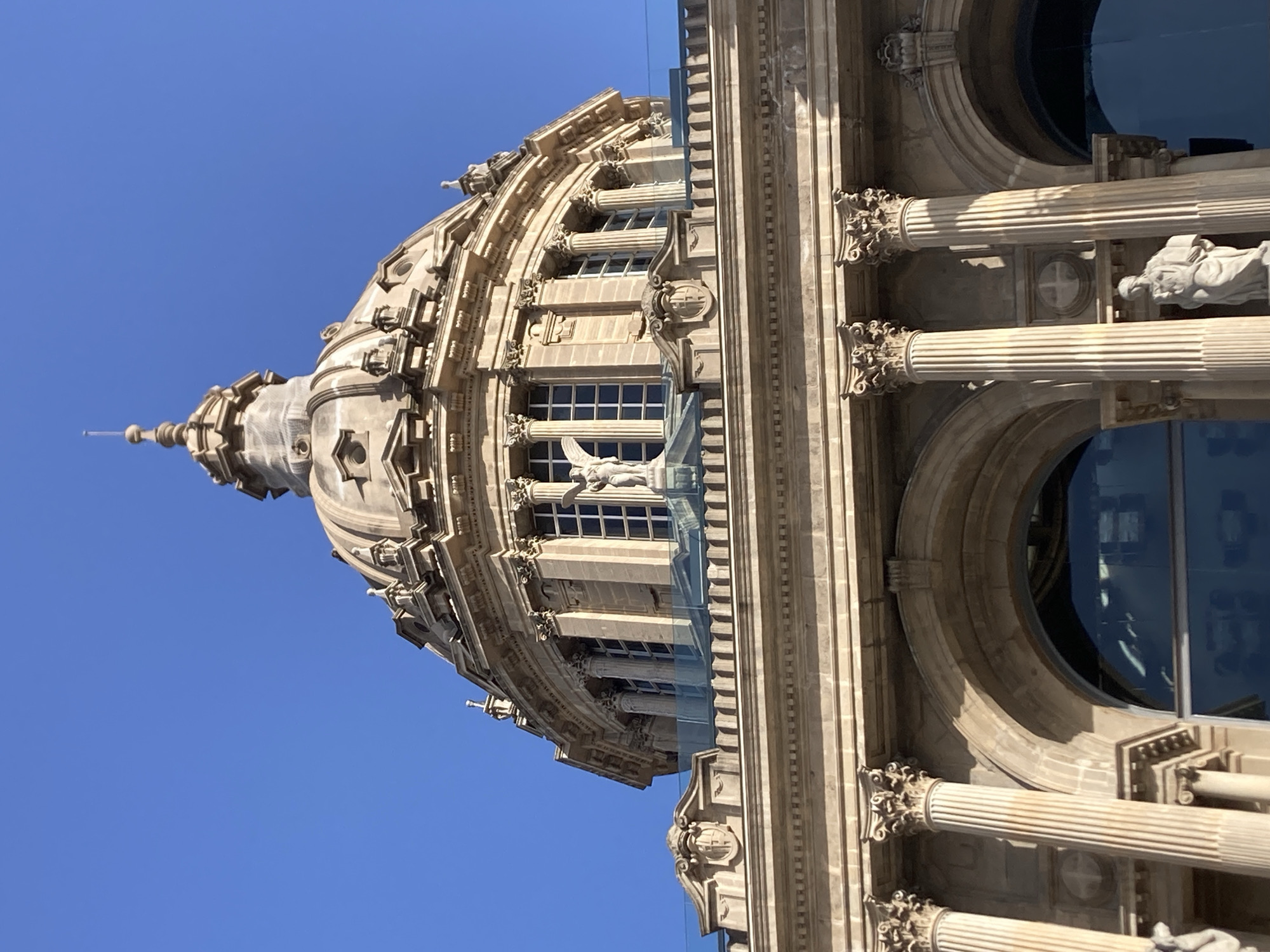 The image shows a close-up of the dome and upper facade of the Palau Nacional, located on Montjuïc hill in Barcelona, Spain. The architecture is neoclassical, characterized by classical columns, ornate detailing, and a prominent dome topped with a spire. A statue of an angelic figure stands prominently on the balcony. The building is constructed of stone and features large, arched windows. The sky is a clear, bright blue.