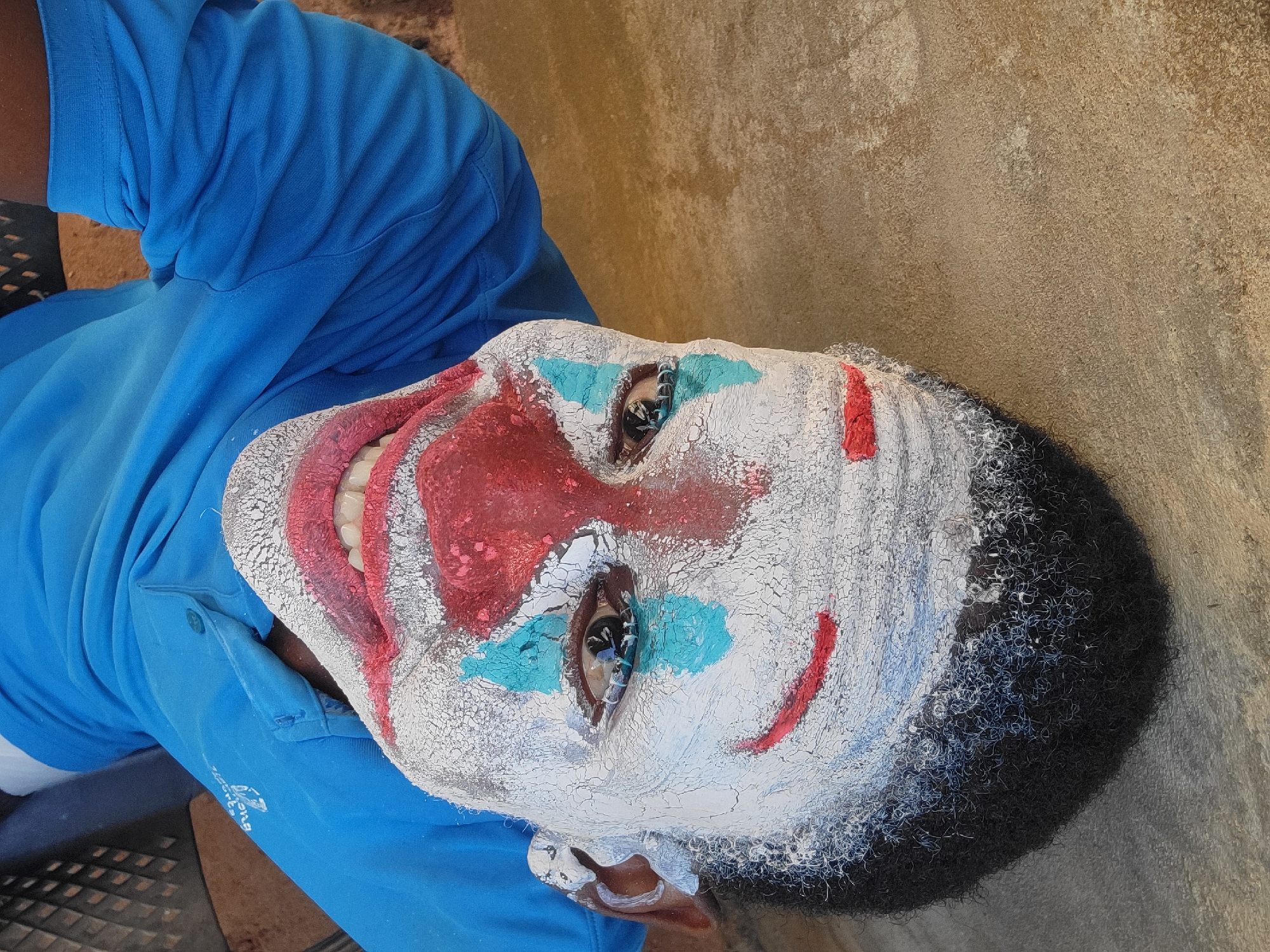 A medium, angled shot captures a person wearing clown makeup. The individual is lying on their back, with their head tilted slightly towards the camera. Their face is covered in white paint, cracked in places, with a large red nose, red markings around the mouth to mimic a smile, and small red streaks on the cheeks. Blue paint is applied around the eyes. The person is wearing a blue polo shirt. The background appears to be an exterior wall, possibly made of concrete or a similar material.
The image was taken in Nkpor, Nigeria.