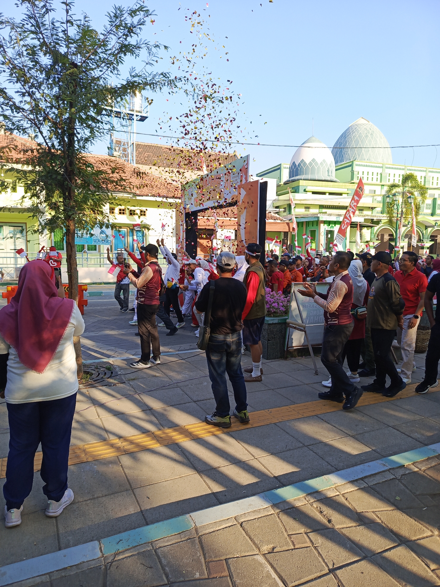 In Semampir, Indonesia, a lively street scene unfolds with a crowd gathered under a clear blue sky. Confetti rains down, adding a festive atmosphere to the event. People, dressed in casual attire, are engaged in various activities, some holding small Indonesian flags, suggesting a patriotic celebration. A group of men in maroon vests and white shirts appear to be part of an organized group, possibly a performance or ceremony. In the background, a mosque with its distinct green domes and architecture stands prominently, indicating the religious and cultural significance of the location. The street is paved with interlocking bricks, and shadows from trees and a nearby structure cast long lines across the ground. The overall impression is one of community gathering and celebration in a vibrant urban setting.