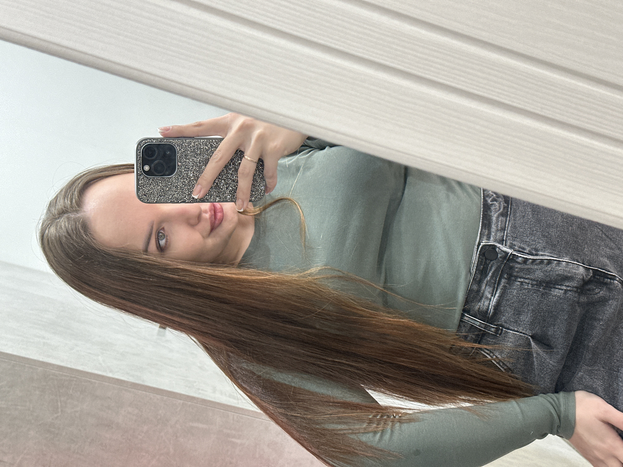 A woman with long brown hair and blue eyes is taking a selfie in front of a mirror. She is holding a phone with a glittery case and wearing a light green long-sleeved shirt and gray jeans. The mirror reflects a white paneled wall. There is no information available to identify the exact location as Chaikovsky, Russia.