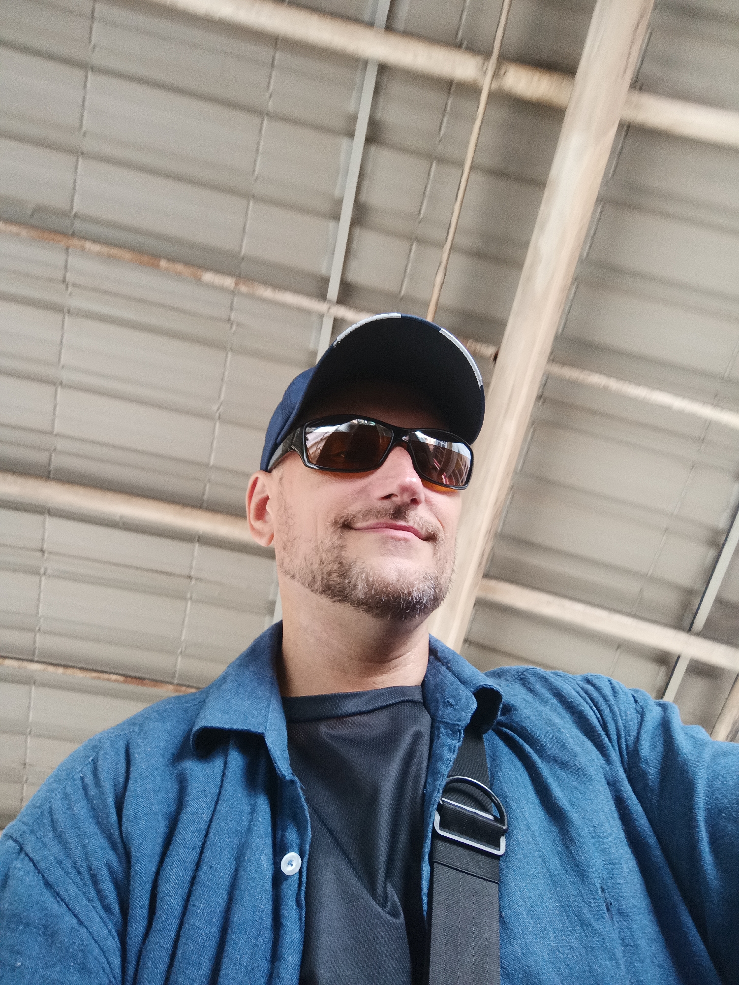The image shows a man wearing a blue denim shirt over a dark t-shirt, a baseball cap, and sunglasses. He has a goatee and is looking at the camera. A black strap with a metal buckle is visible across his chest. The background appears to be an industrial or warehouse-like structure with a corrugated metal roof and exposed beams. The lighting suggests an indoor setting, possibly during the daytime. The image was taken in Kuala Lumpur, Malaysia.