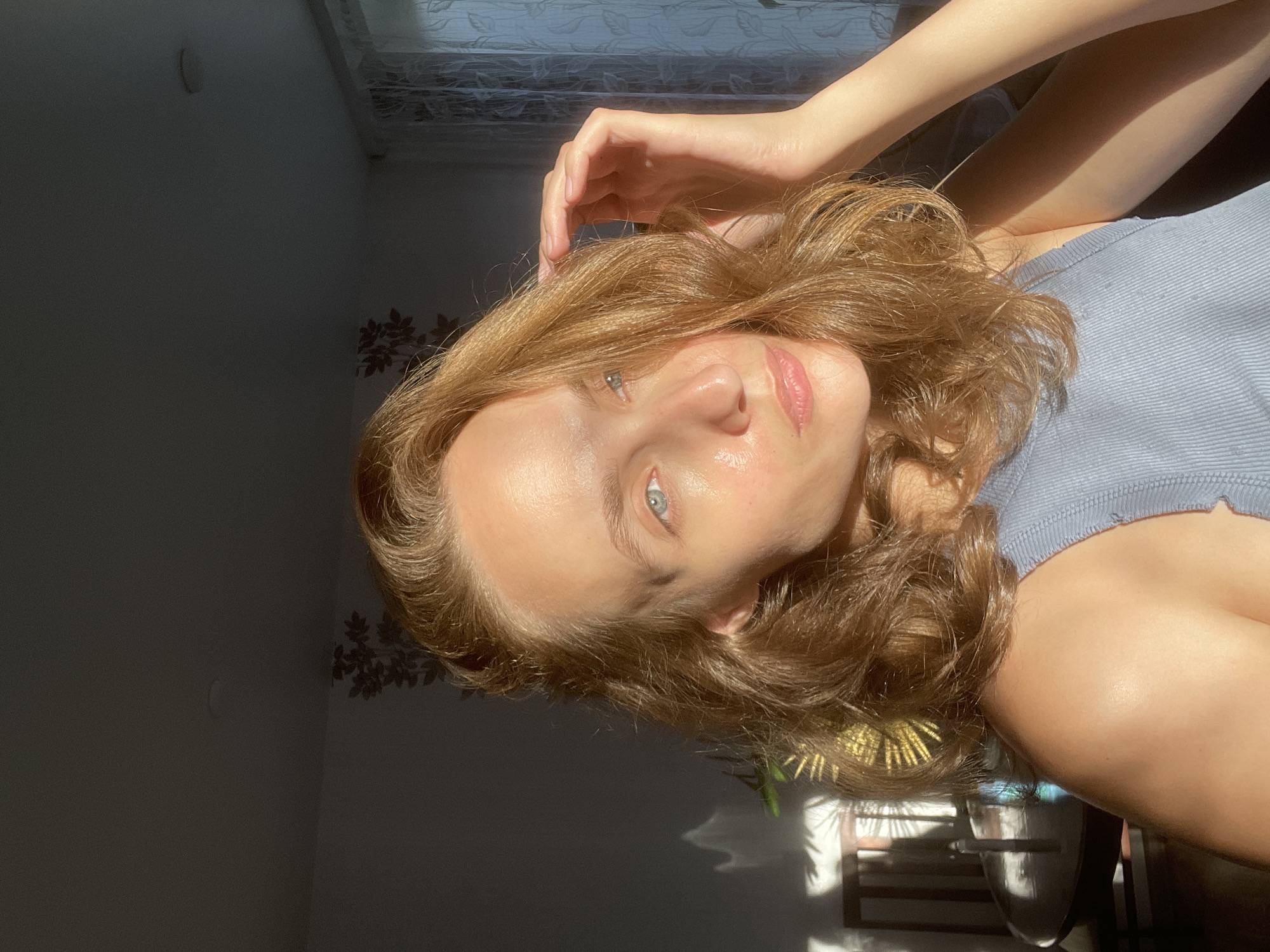 A young woman is depicted in a close-up, brightly lit portrait, presumably taken indoors in Sharkan, Russia. She looks directly at the camera with a calm, natural expression, her head tilted slightly. Her light brown, wavy hair frames her face, which is illuminated by strong, direct sunlight. She has fair skin, light blue-grey eyes, and her lips appear glossy. She is wearing a light blue or grey ribbed tank top. Her right hand is visible, positioned near her head, lightly touching her hair, while her left arm is bent, showing a tanned forearm.
The lighting is a prominent feature, with harsh sunlight creating distinct highlights on her skin and hair, and casting sharp shadows in the background. The scene suggests it is either morning or late afternoon. The background is partially obscured by shadows, revealing a dark wall on the left with a subtle, dark floral or leaf pattern. To the right and behind her, elements such as a textured curtain or window are visible, along with glimpses of indoor plants, one appearing to have golden, spiky foliage. The overall mood is serene and natural. No visible text is present in the image.