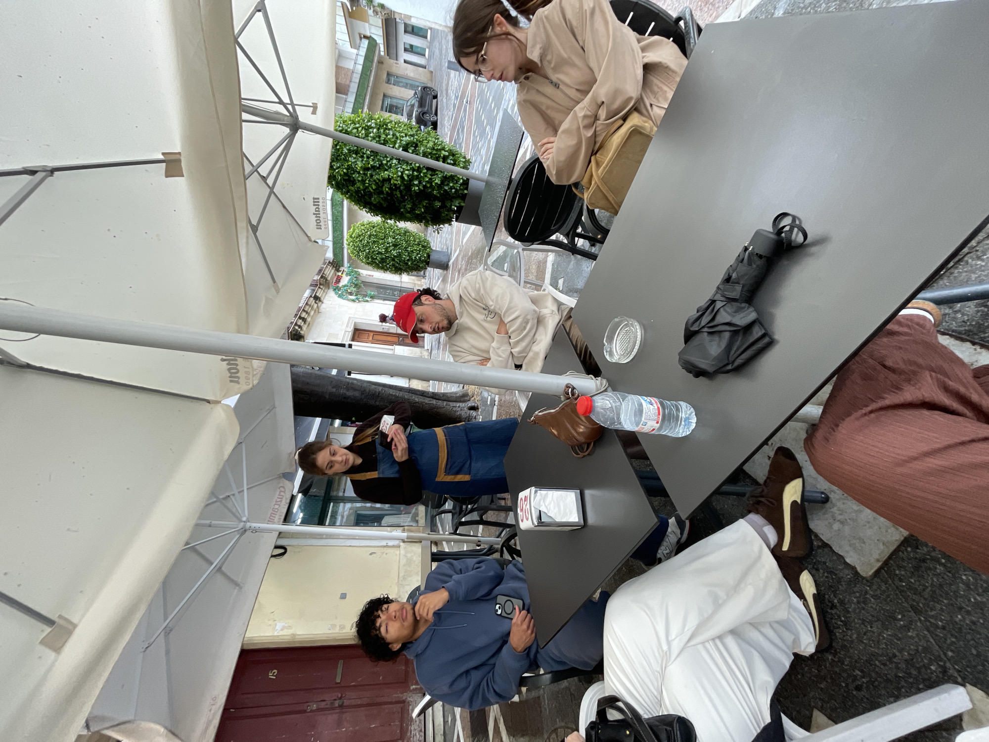 The scene depicts a group of people at an outdoor cafe, likely located in a pedestrian area with shops. A couple sits at a table, one wearing a tan jacket and glasses, the other a white shirt and red baseball cap. A waiter wearing a denim apron and black shirt is standing nearby with what appears to be a phone. At another table, a person wearing a blue hoodie is holding a phone, while another person is wearing white pants and brown shoes with white soles. On the tables are items such as a water bottle, a black umbrella, a purse, a tissue box, and a glass ashtray. The scene is sheltered by white umbrellas. The environment appears to be a street in the city of Jerez De La Frontera, Spain.