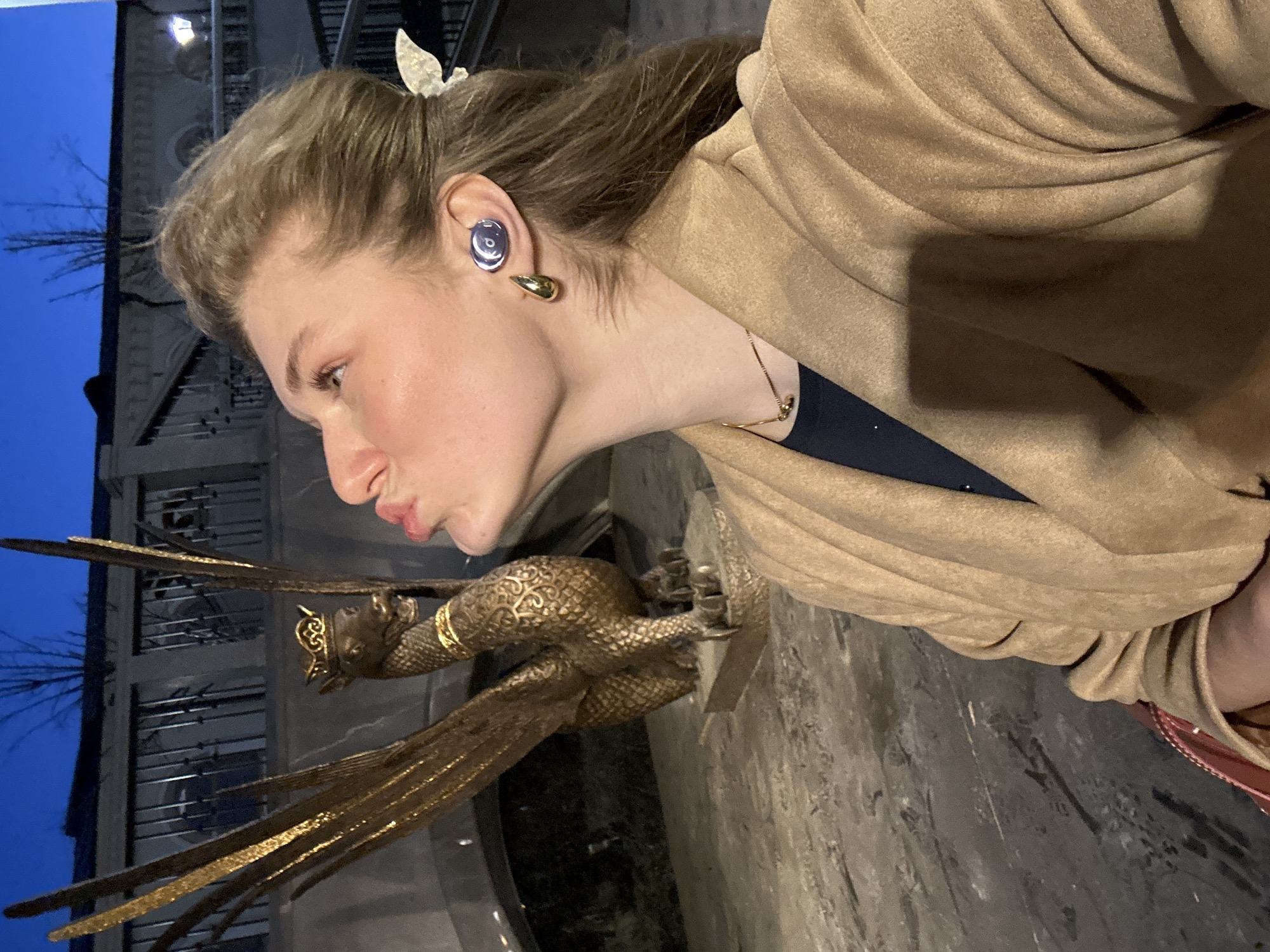 A woman stands next to a golden griffin statue in Kazan, Russia. She is looking at the statue with her lips pursed. She is wearing a tan coat, a gold necklace, and earrings, and her hair is partially clipped back with a butterfly clip. The statue is a winged griffin with intricate detailing. The background shows a building at dusk.