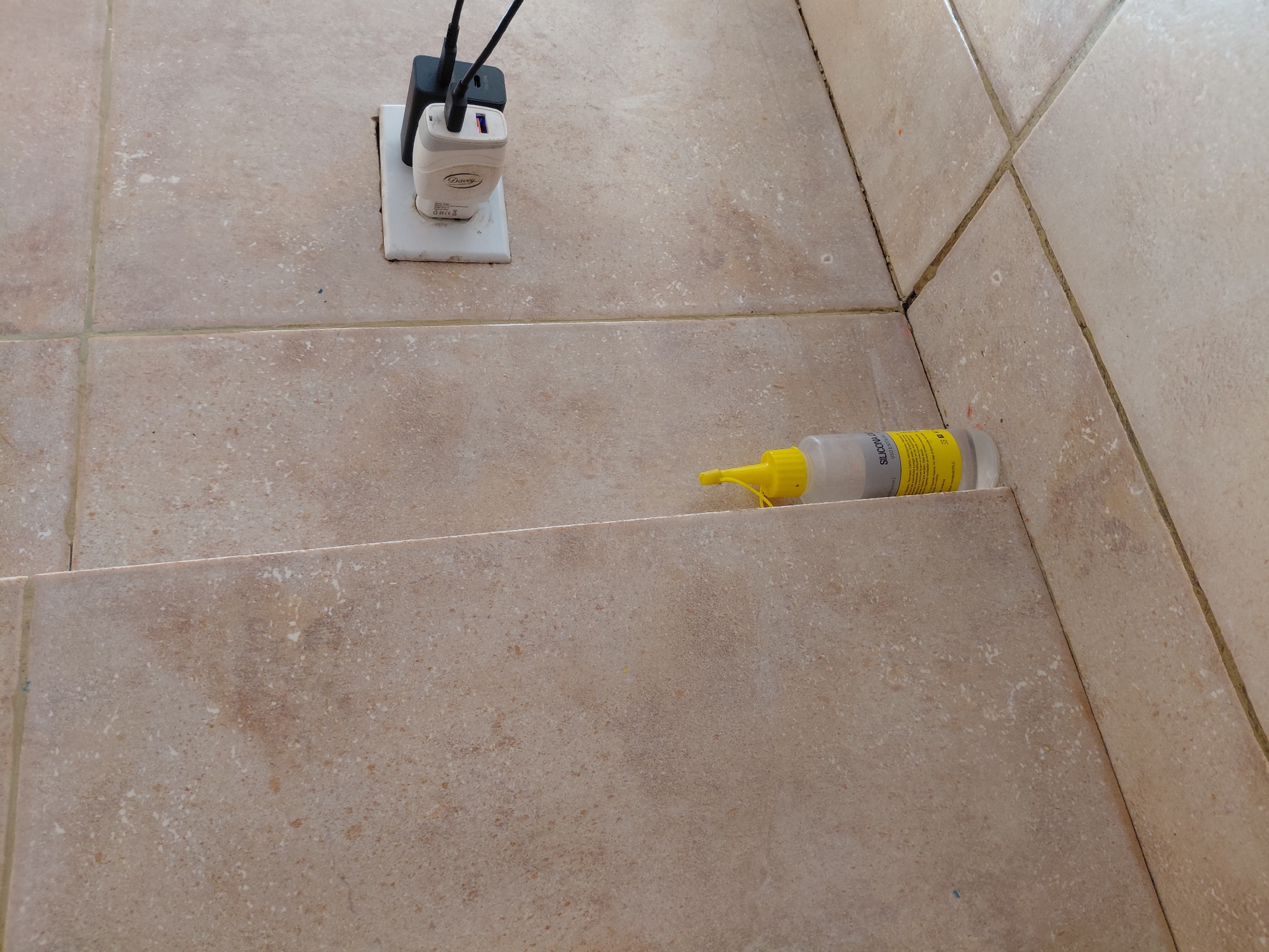 The image presents an indoor scene, likely a floor or wall junction, composed of light brown/beige speckled ceramic tiles separated by dark gray grout lines. In the upper left quadrant, a white electrical outlet is mounted on the tiled surface. A black charger and a white charger, branded "Davey," are plugged into the outlet, with black cables extending upwards from them.
Towards the right side and lower in the frame, a clear plastic bottle with a bright yellow cap and nozzle lies horizontally on the tiles. The bottle's label clearly displays "SILICONA" and "Silicona Multiuso," indicating it contains multi-purpose silicone, with "30ml" also visible, denoting its volume.
The setting appears to be a functional area, possibly a utility corner or a section of a room. The bright, even lighting suggests it is daytime. There are no people visible, and no specific activity is taking place; the objects appear to be static. The presence of chargers implies electronic devices are or were being powered, while the silicone bottle suggests recent or impending sealing or repair work. This scene could be from a home or commercial building in Santa Ana, Venezuela, reflecting typical daily items and materials.