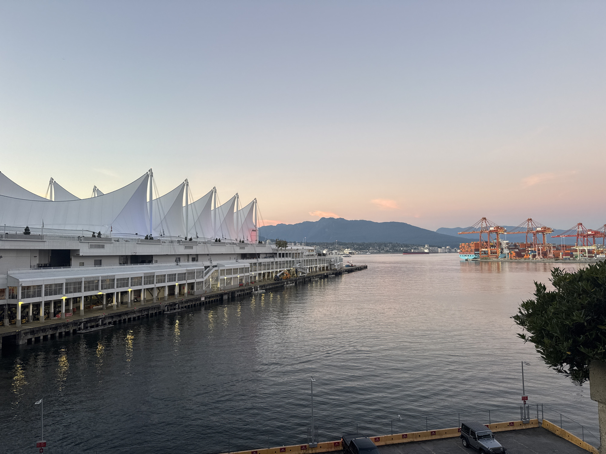The image depicts a waterfront scene in Vancouver, Canada, likely at Canada Place. In the foreground, a body of water reflects the soft colors of the sky and the lights from the buildings. On the left, the distinctive white sail-like structures of Canada Place are visible, with a long, multi-story building extending along the waterfront. Lights are illuminated along the lower level of this building, casting reflections on the water. Further out in the harbor, a large cargo ship filled with containers is docked, surrounded by multiple towering orange cranes. The background features a range of mountains under a pale sky tinged with sunset colors. The overall scene suggests an early evening ambiance in a busy port city.