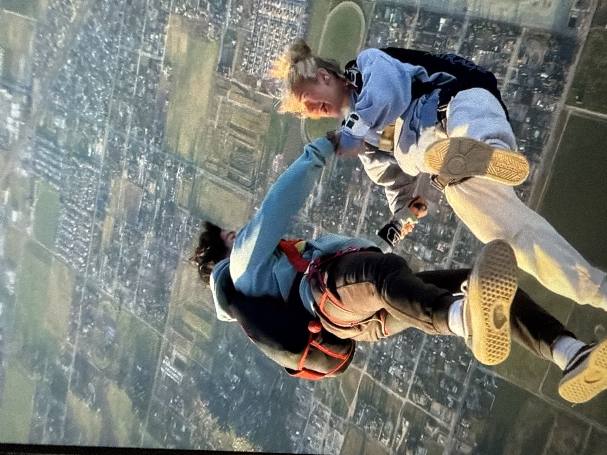 Two individuals are skydiving, holding hands. The person on the left wears a light blue hooded jacket, dark pants, and a red-accented harness. The person on the right has blonde hair in a bun, a light blue and black jacket, and white pants. The ground below shows a cityscape with patches of green. A circular track can be seen among the buildings. Location: Unknown, Unknown.