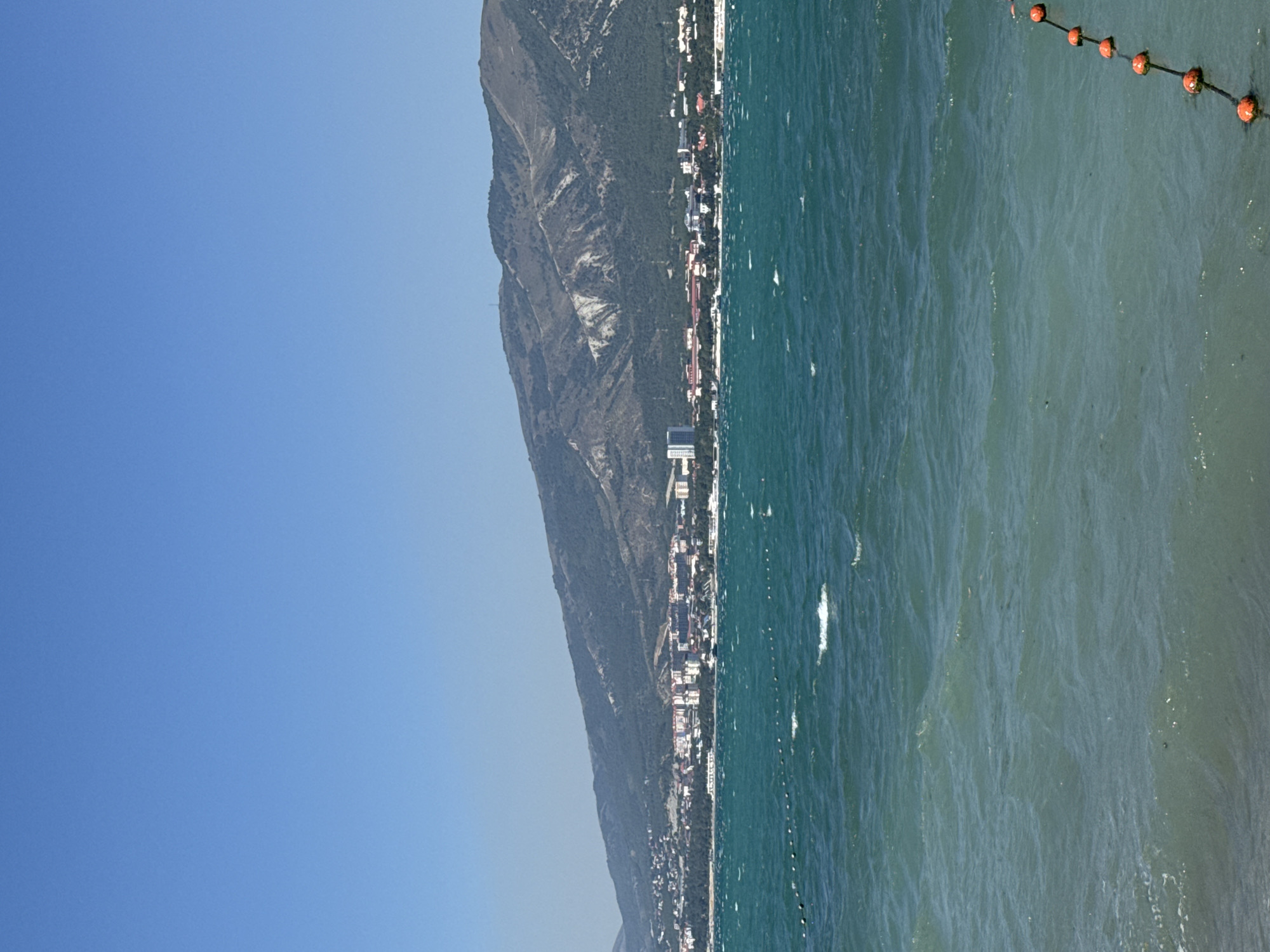 The image depicts a seaside view of Gelendzhik, Russia, with a clear blue sky dominating the upper portion of the frame. A coastal city with low-rise buildings and a few taller structures is visible along the shoreline. Behind the city, a large, verdant mountain range slopes upwards, its peak partially obscured by haze. The foreground is filled with the turquoise waters of the sea, characterized by moderate waves and whitecaps, indicating windy conditions. A line of orange buoys is visible in the water, demarcating an area. The overall scene is tranquil, with no visible people or animals. The lighting suggests it is daytime, likely late morning or afternoon, with the sun casting a natural light on the landscape.