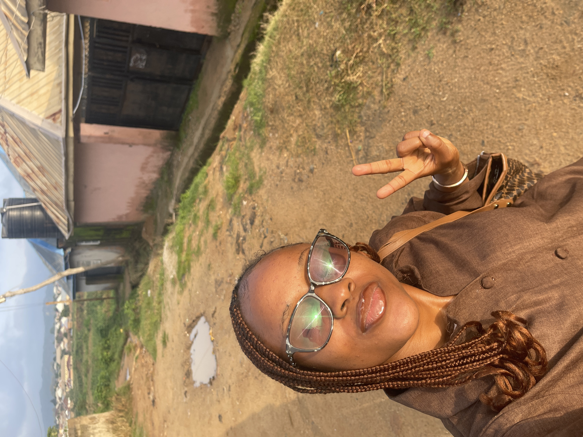 A young woman is taking a selfie outdoors, with her head tilted back and a peace sign displayed. She is wearing a brown shirt and glasses. Her hair is braided and hangs down her back. She has on lip gloss. The background shows a dirt path with some patchy grass and a puddle of water. In the distance, there are buildings and a hillside covered with more structures. The lighting suggests it is daytime, likely late afternoon, with the sun casting reflections on her glasses. The scene appears to be in a rural or semi-urban area, possibly Rafin Par, Nigeria. The woman's expression seems joyful and relaxed. There is no visible text in the image.
