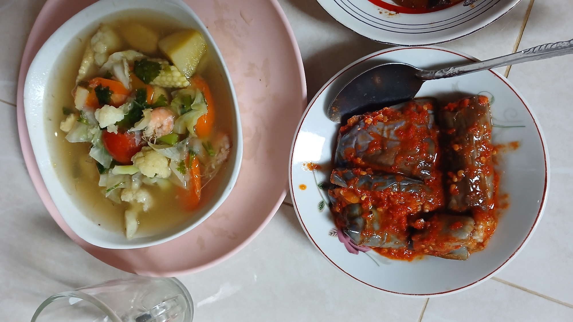 A top-down shot reveals two dishes on a white-tiled surface. On the left, a white bowl sits on a pink plate, containing a clear soup filled with mixed vegetables, including cauliflower, cabbage, carrots, tomatoes, and corn. Some shrimp are visible in the soup. To the right, a white plate with a floral pattern holds sliced eggplant covered in a red chili sauce, with a metal spoon lying on top. A partially visible drinking glass is in the bottom left corner, and a bit of another plate is visible in the upper right. There is no indication of the location being Lhokseumawe, Indonesia.