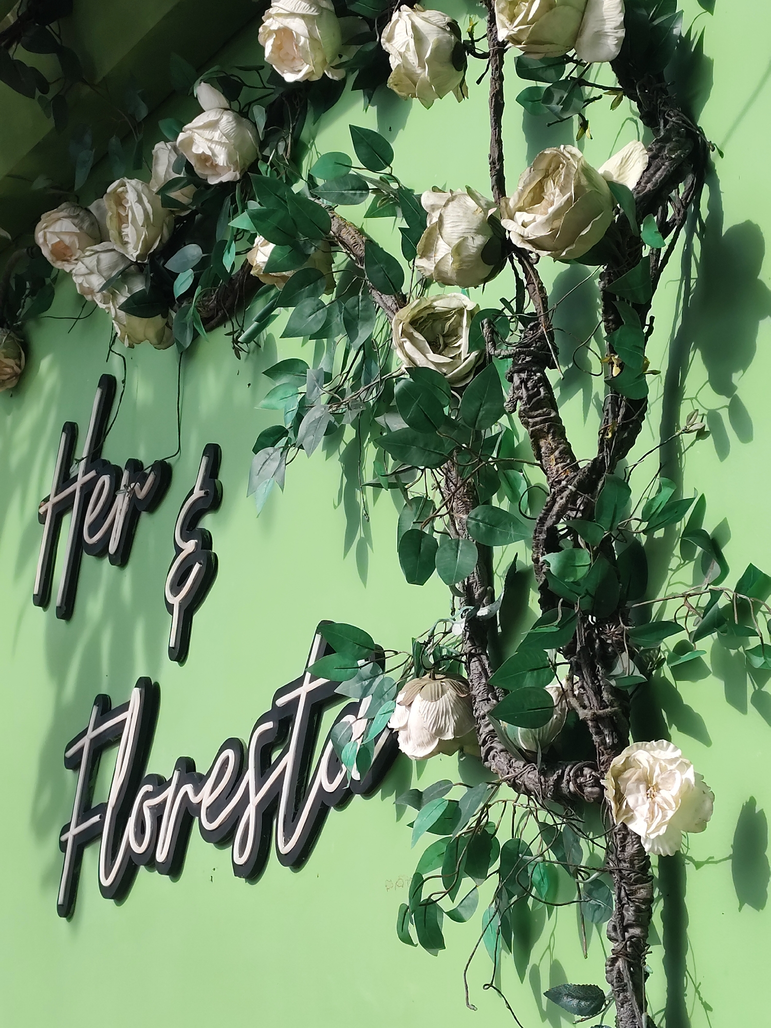 An outdoor scene features artificial floral decorations and signage against a light green wall. The floral arrangement consists of cream-colored roses and dark green leaves intertwined with a dark brown, twisted vine. The vines climb vertically, with some branches extending horizontally. The signage, which appears to be neon or LED letters, is mounted on the wall. The top line of text reads "Her &" in a script font. The second line of text, positioned below the first, reads "Floresta" in a similar script font. Both lines of text have a dark outer layer and a lighter inner glow, suggesting they are illuminated. The wall behind the decorations casts shadows from the vines and leaves. The overall scene suggests a decorative element for a shop or establishment, possibly an entrance or facade. The location is identified as Da Lat, Vietnam, a city known for its floral attractions and French colonial architecture, which might explain the style of decoration. No people are visible in the image.