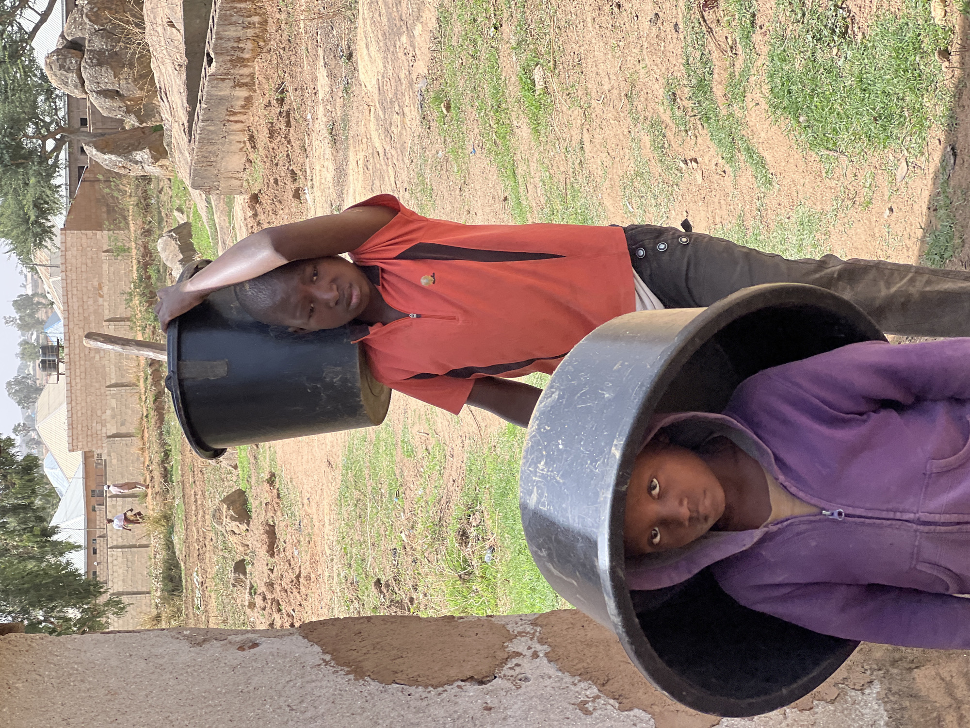 Two children pose with large, black plastic containers in an outdoor setting in Bukuru, Nigeria. One child, wearing a red shirt with black accents and dark pants, rests his head on the rim of a container positioned to his left. The other child is inside a container in a purple hooded sweatshirt, looking directly at the camera. The background consists of dry, grassy terrain, a stone structure, and buildings in the distance.