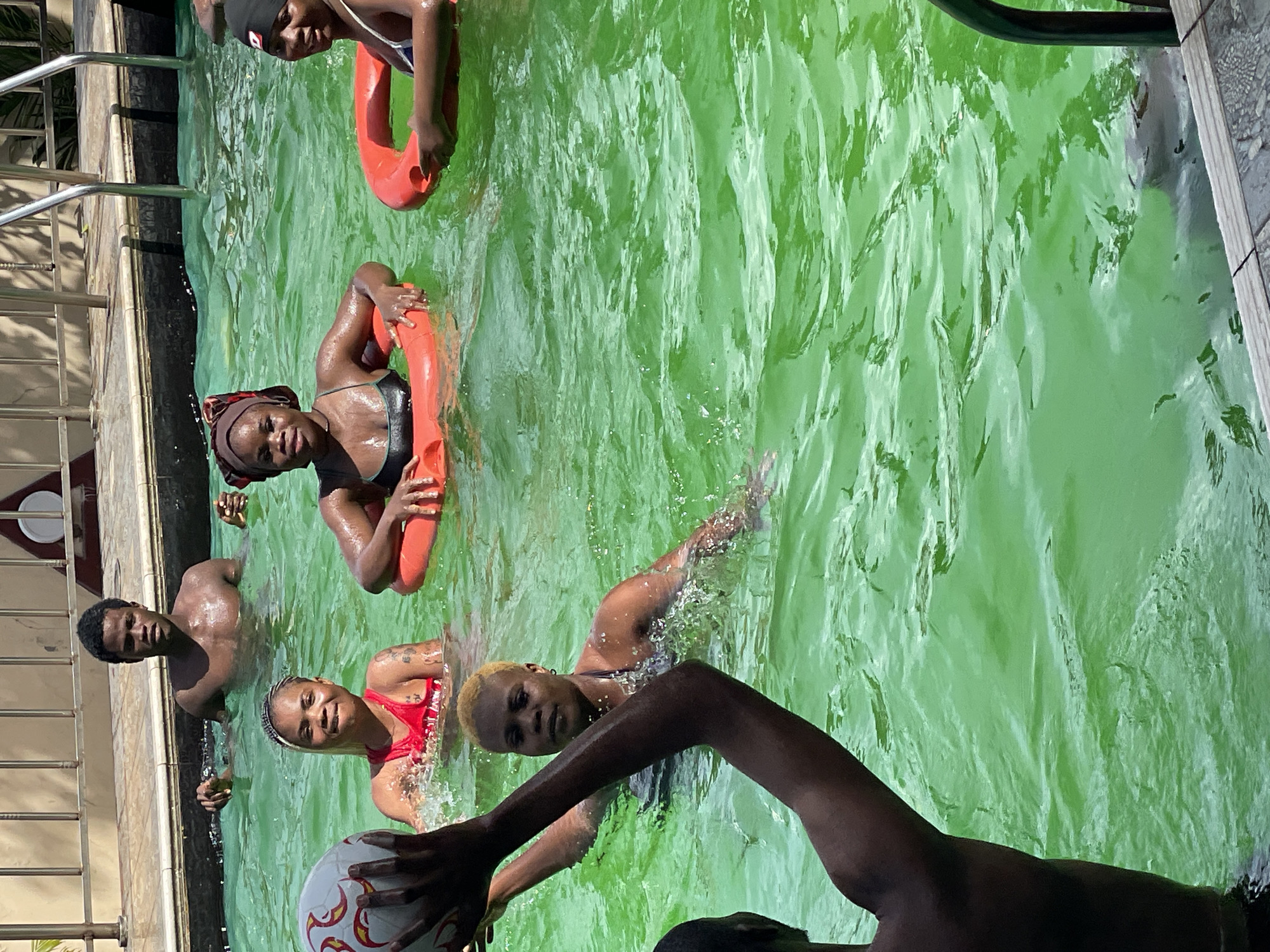 In an outdoor swimming pool filled with bright green water, a group of young adults are enjoying a sunny day. A young man with dark skin and short hair is partially submerged in the water, looking towards the camera. Beside him, a young woman with blonde hair, wearing a red swimsuit, is splashing in the water and holding a white and red patterned ball. To her right, another young woman, wearing a dark bikini, is holding a bright orange inflatable ring. Further to the right, a young woman with dark skin and a dark headscarf is also holding an orange inflatable ring. The pool has a concrete edge and is surrounded by a metal fence. The lighting suggests it is daytime with clear weather.