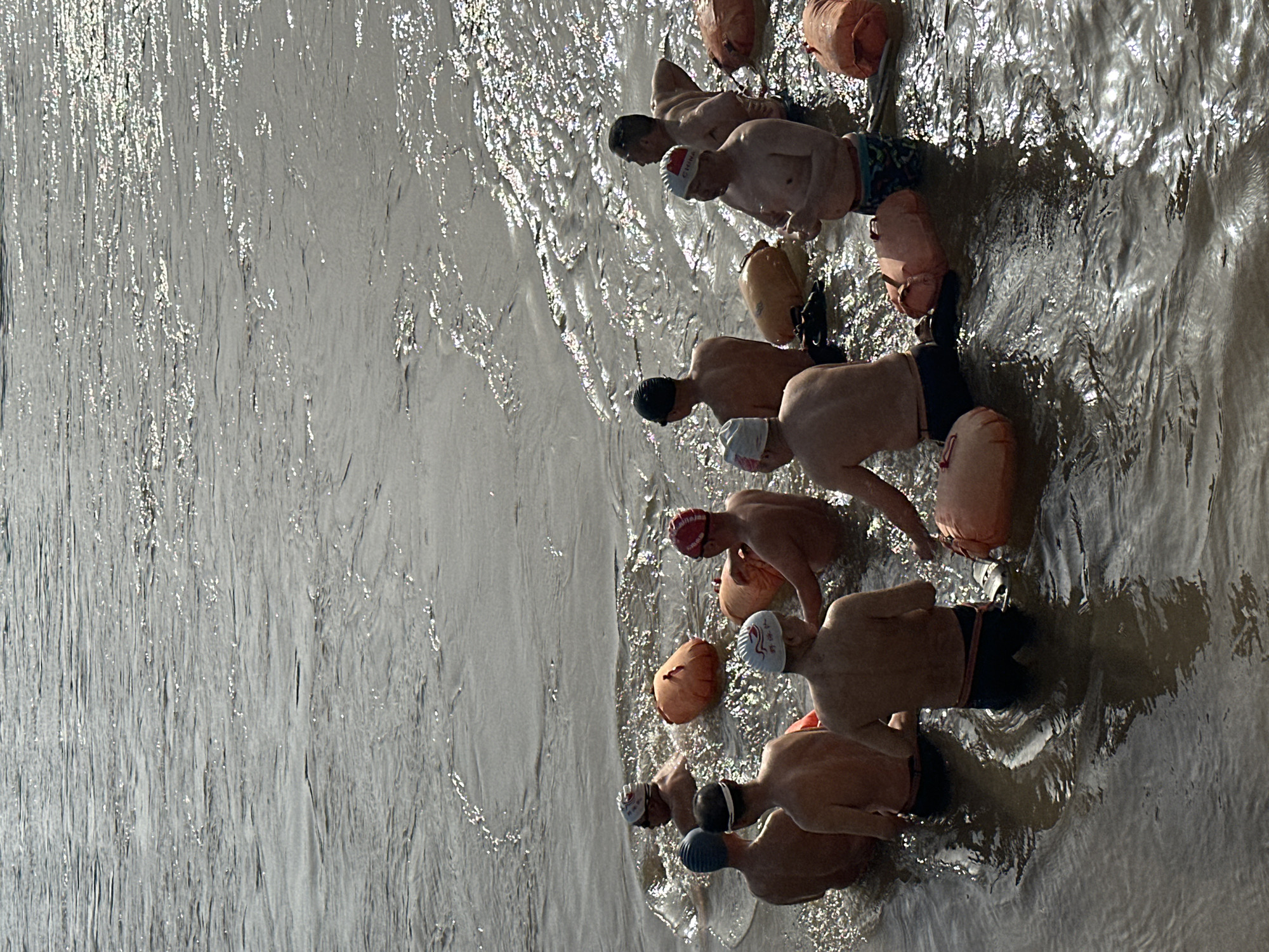 The image, oriented vertically, captures a group of people standing partially submerged in a body of water, likely a river, under bright sunlight. There are at least seven individuals visible, all appearing to be adult men. They are dressed in swimming trunks or similar attire, and each wears a swimming cap. Several orange inflatable swim buoys are visible, some attached to the swimmers, suggesting a safety measure for open-water swimming.
The activity taking place appears to be an open-water swimming event or group training session, with the participants either preparing to swim or taking a break. The water surface is highly reflective, with strong glints of sunlight indicating a sunny day. The water itself has a grayish-brown hue, characteristic of many large rivers.
Notable details include the brilliant reflections on the water, suggesting either early morning or late afternoon light. On one white swimming cap, the word "CHINA" is clearly visible in red lettering. Another red swim cap also has "CHINA" written on it in white. The water's texture shows ripples and slight currents. The group seems to be positioned along a shoreline or shallow edge of the river. Given the location context of Jiangjin, China, it is highly probable that the scene takes place in the Yangtze River. The overall impression is one of a communal and perhaps competitive or recreational aquatic activity.