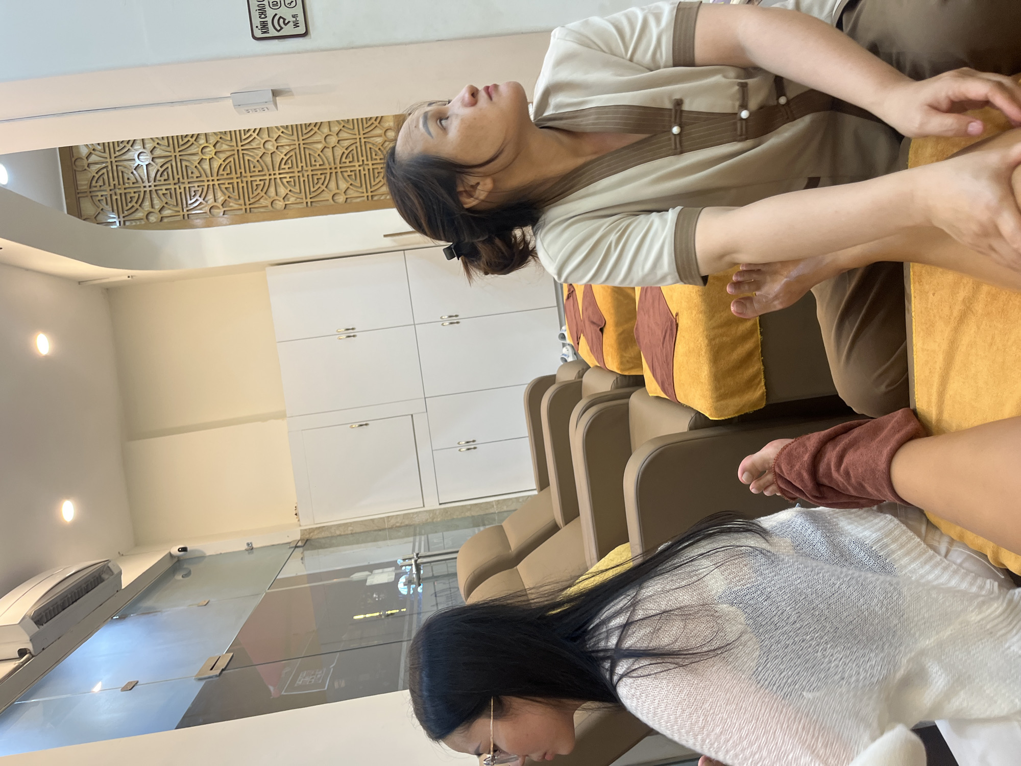 The image depicts two women in what appears to be a massage parlor in Nha Trang Ward, Vietnam. One woman, wearing glasses and a light-colored knitted sweater, is seated on a massage chair with her legs extended forward, partially covered by a dark reddish-brown towel. The second woman, wearing a beige uniform with brown trim and pearl-button closures, is also seated and appears to be performing a massage on the first woman's leg. The background reveals rows of similar massage chairs, some covered with yellow and brown fabric. White cabinets with double doors are visible on the wall, and a decorative wooden panel with an intricate geometric pattern is also present. A sign with Vietnamese text and Wi-Fi symbols is mounted on the wall to the right. An air conditioning unit is mounted on the ceiling, and a security camera is visible on the upper left wall.