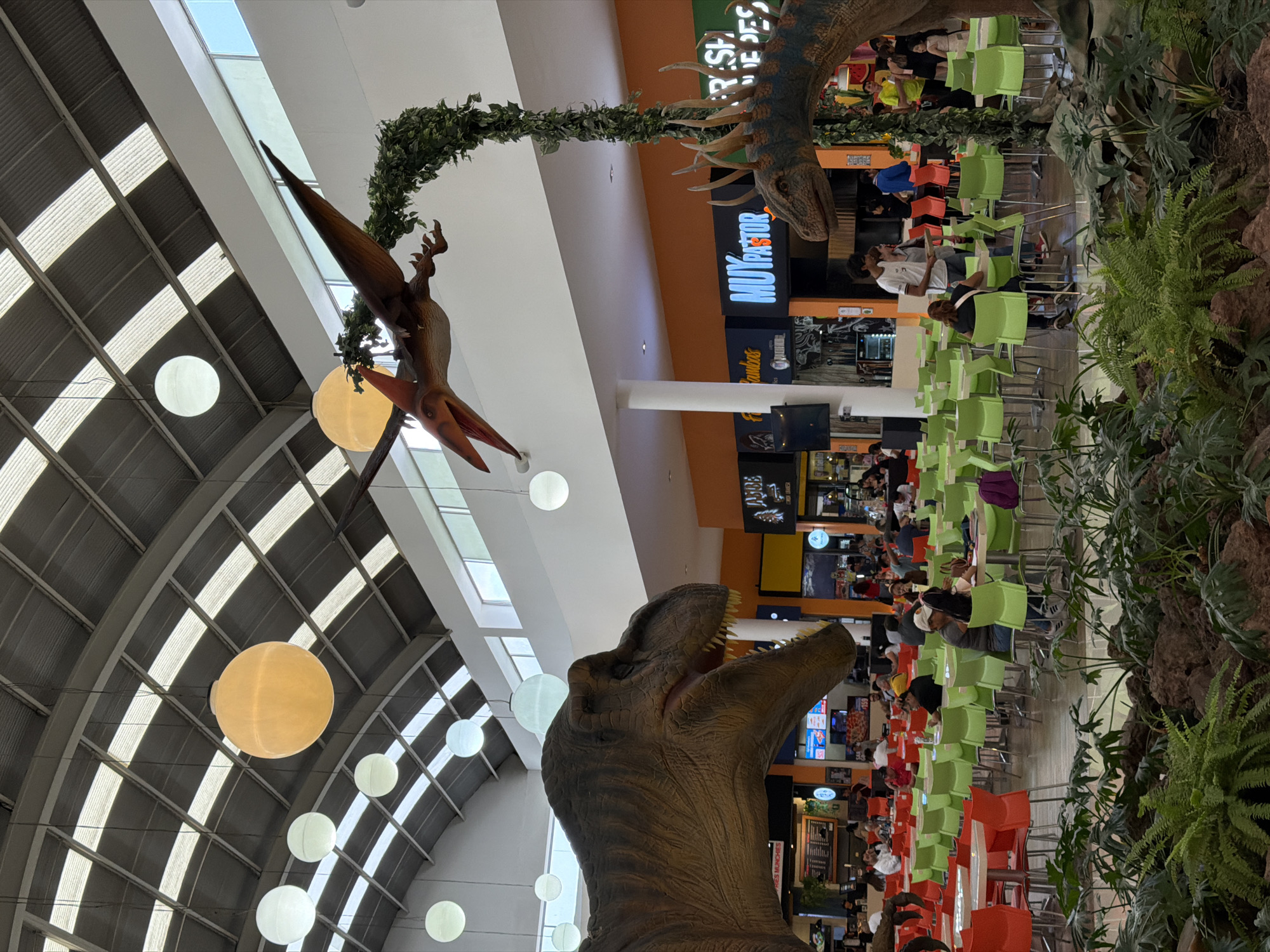 The image shows the interior of a food court featuring animatronic dinosaurs suspended from the ceiling. A Tyrannosaurus Rex is in the foreground, while a Pterodactyl and another dinosaur are visible in the background. The food court has numerous tables and chairs occupied by people eating. Various restaurant signs are also visible. The ceiling structure has an arched design with spherical light fixtures. Lush green plants are arranged as decoration around the seating area. The location is in San Francisco Ocotlán (Ocotlán), Mexico.