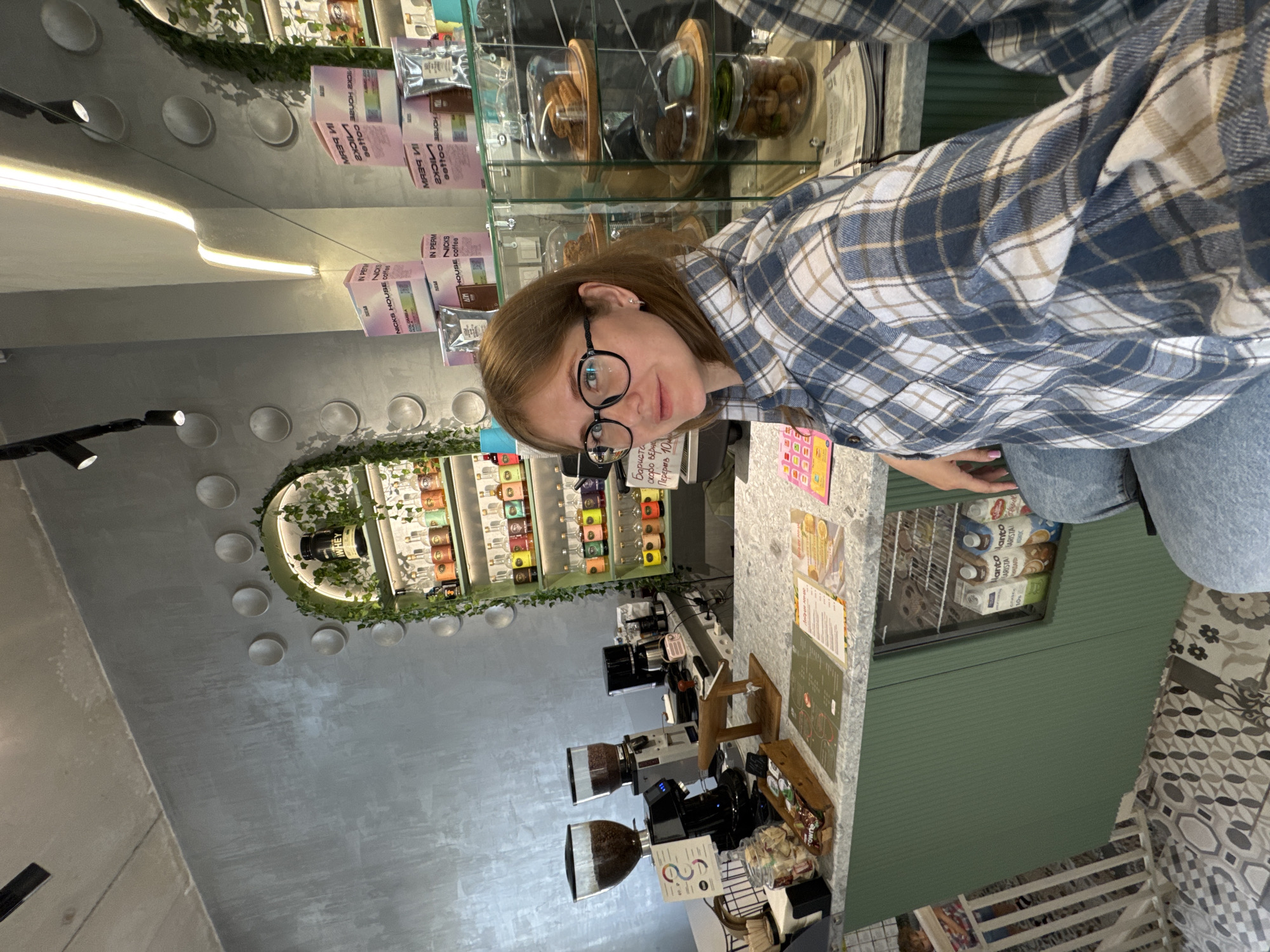 In a brightly lit, modern cafe located in the city of Perm, Russia, a young woman with blonde hair and glasses sits at a counter, facing the camera. She is wearing a blue and white plaid shirt and blue jeans. Behind her, the cafe's interior is visible, featuring a green counter with a speckled surface, coffee grinders, a coffee machine, and shelves stocked with bottles and boxes. A glass display case showcases baked goods. The walls are a textured gray, and a decorative archway with greenery and lights adds to the ambiance. The overall scene captures a casual moment in a contemporary coffee shop setting.
