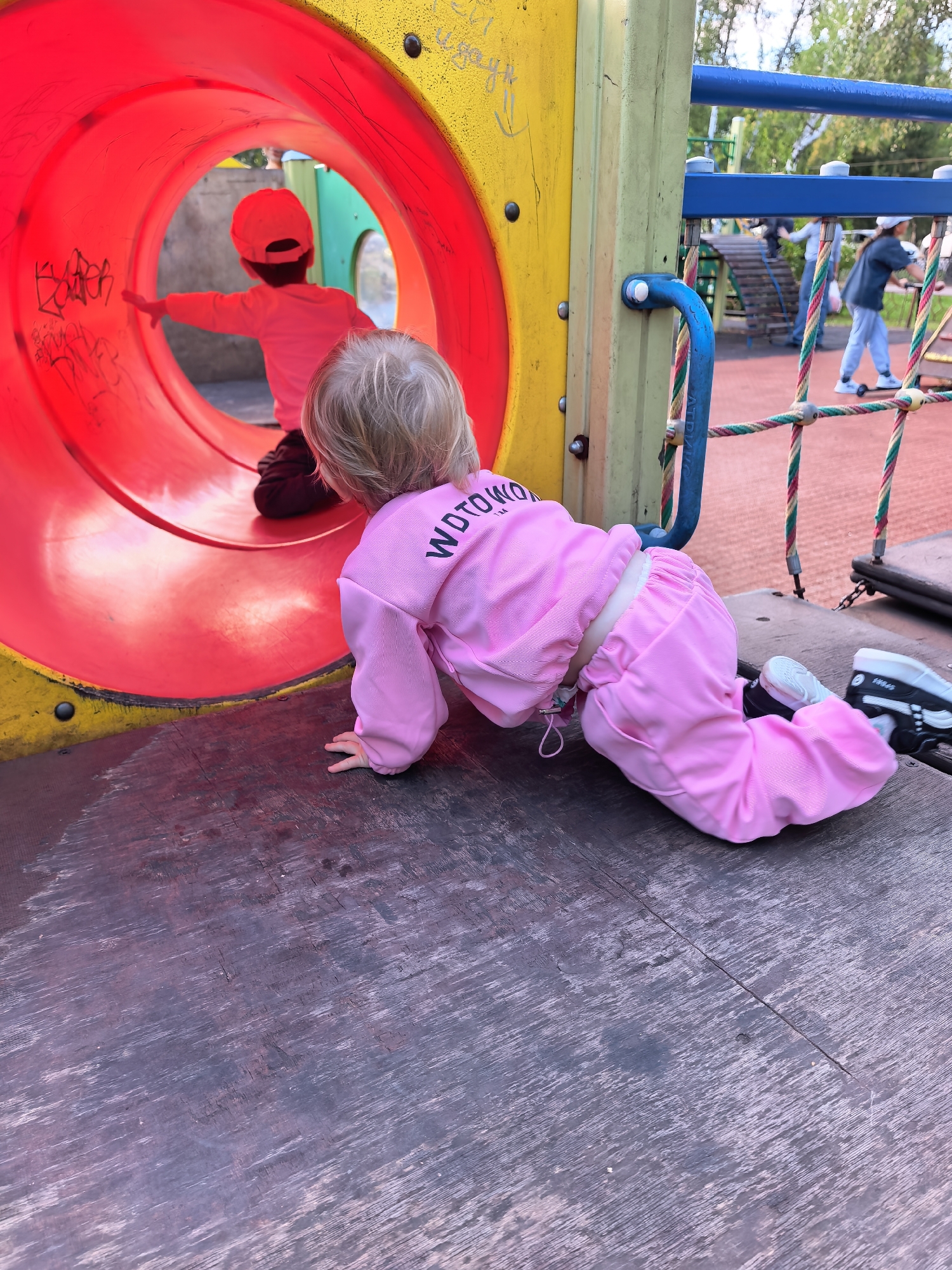 A blonde toddler wearing a pink tracksuit and sneakers is crawling on a wooden surface at a playground. The toddler is facing away from the camera, looking into a red tunnel slide. Inside the tunnel, a child wearing a red cap and a pink shirt is visible. The playground features yellow and green structures, blue railings, and a red net climbing structure in the background. In the distance, other children are playing. The environment appears to be an outdoor park with trees and a sky visible. The image was taken in the city of Sterlitamak, Russia.