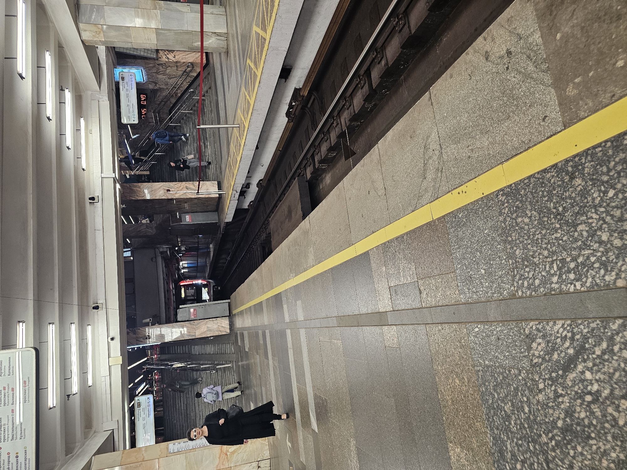 A woman wearing a long black coat and sunglasses walks along a subway platform in Moscow, Russia. She carries a black shoulder bag. In the background, several people ascend a wide set of stairs, and a subway train is visible on the tracks to the right. The platform floor is tiled with a patterned granite, and a yellow line marks the edge of the platform. Overhead, fluorescent lights illuminate the station. Signs in Cyrillic script are visible on the station walls and ceiling.