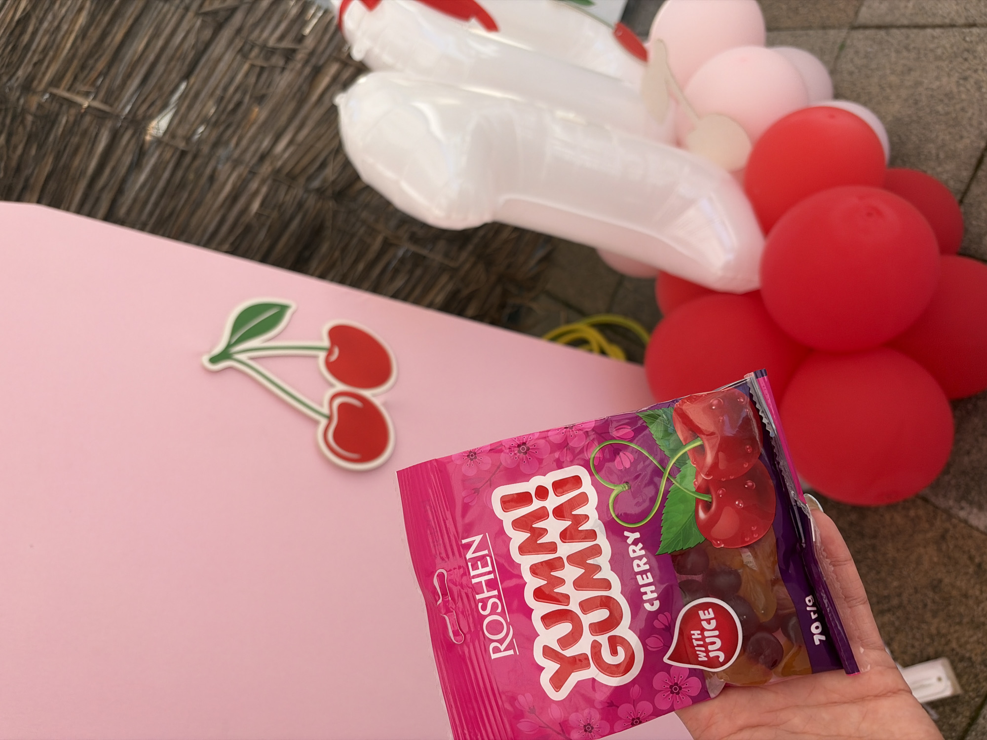 A hand holds a pink bag of Roshen Yummy Gummi Cherry flavored candies. The bag has a cherry design and text indicating "with juice" and "70 g/r". In the background, a pink surface with a cherry decal is visible, along with a cluster of red and pink balloons. The scene appears to be set outdoors, possibly in Kolarovo, Bulgaria, given the cobblestone-like ground texture and the visible outdoor elements.