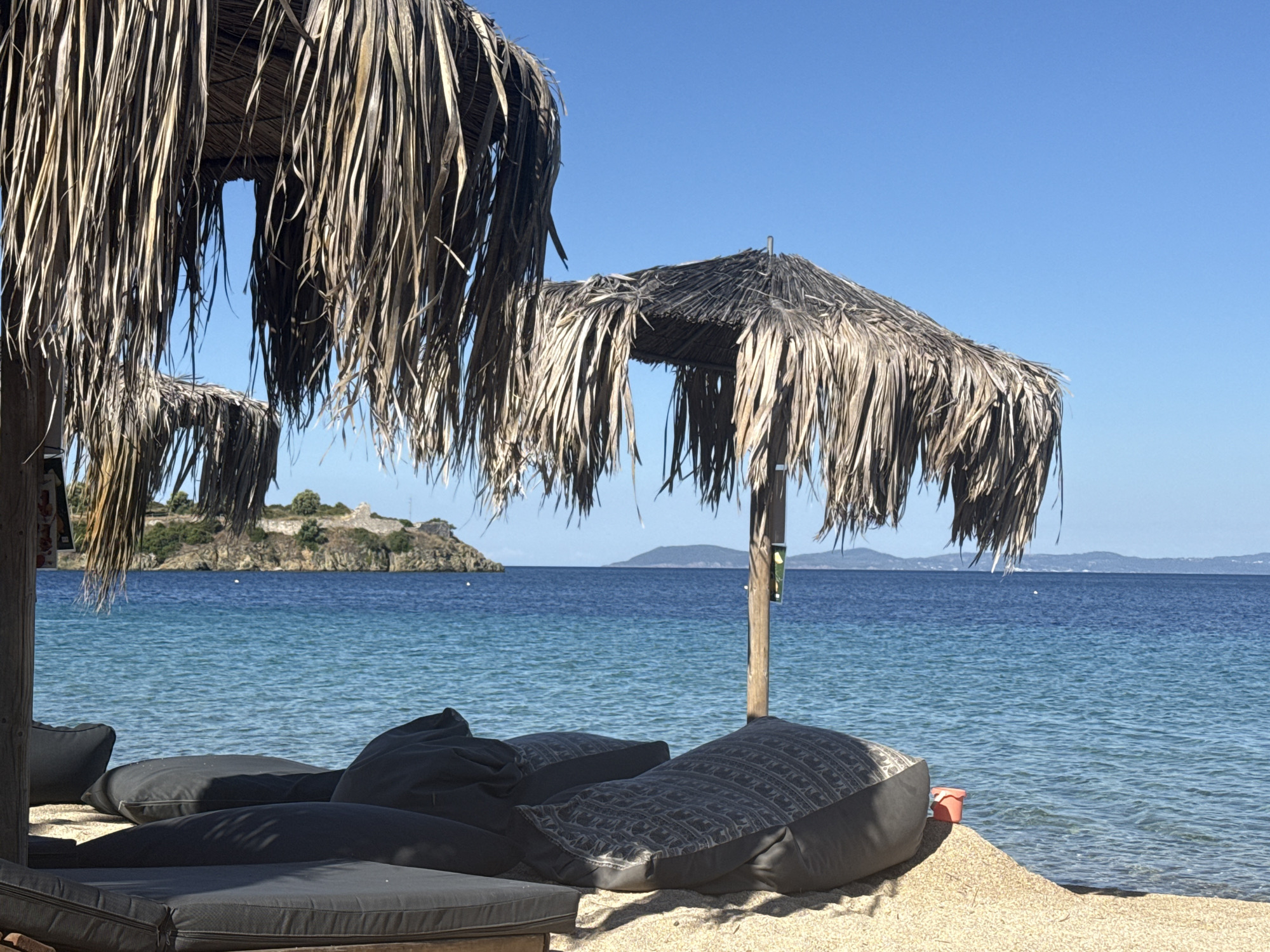 This is a photograph of a serene beach scene in Toroni, Greece, under a clear blue sky. The foreground is occupied by comfortable-looking lounge seating, including cushions and beanbag chairs, on a sandy beach. Two thatched umbrellas provide shade over the seating area. In the middle ground, the calm, turquoise waters of the Aegean Sea gently lap the shore. Across the water, in the background, a rocky island with ancient ruins, possibly a castle, is visible. Further in the distance, rolling hills and mountains form the coastline. The overall impression is one of tranquility and relaxation, characteristic of a Greek beach resort.