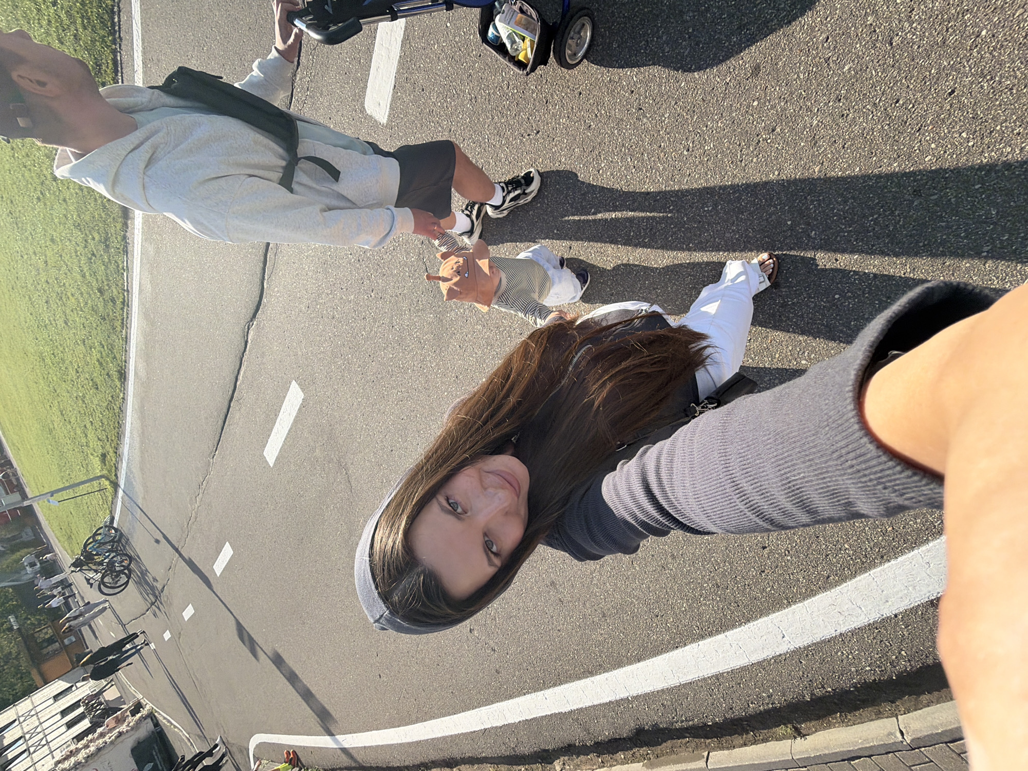 In Odintsovo, Russia, a woman with long brown hair and wearing a grey hooded jacket takes a selfie from a low angle. She is walking on an asphalt path with white lane markings, possibly a bike path or sidewalk, adjacent to a grassy area and buildings. To her right, a man in a light grey hoodie and dark shorts walks, pushing a stroller that contains items like bottles and a bag. A small child, wearing a striped shirt and a light brown hat with ear-like decorations, walks alongside the man, holding his hand. In the background, other people are visible walking, and two bicycles are parked. The sun is casting long shadows across the path, indicating it is likely late afternoon. The overall scene appears to be a public space within the city, possibly a park or recreational area.