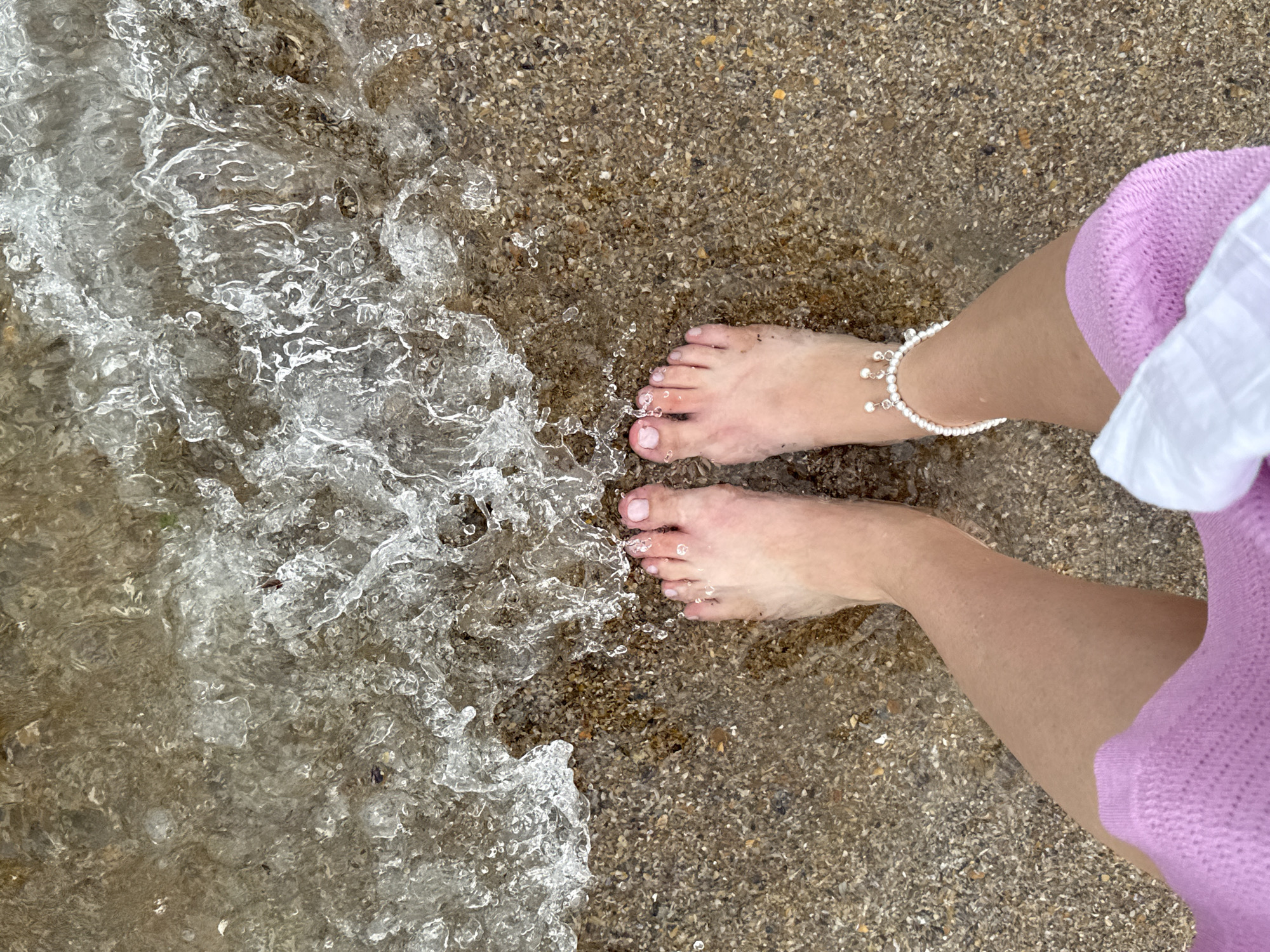 A first-person perspective shot from above shows a person's bare feet standing on a pebbled shore where shallow, clear water is lapping. The person is wearing a pink knitted garment on their legs and a white accessory that wraps around their lower leg. A pearl anklet is visible on the right ankle. The scene appears to be a beach, possibly in Derbent, Russia, given the context. The water is creating frothy waves as it washes over the pebbles.