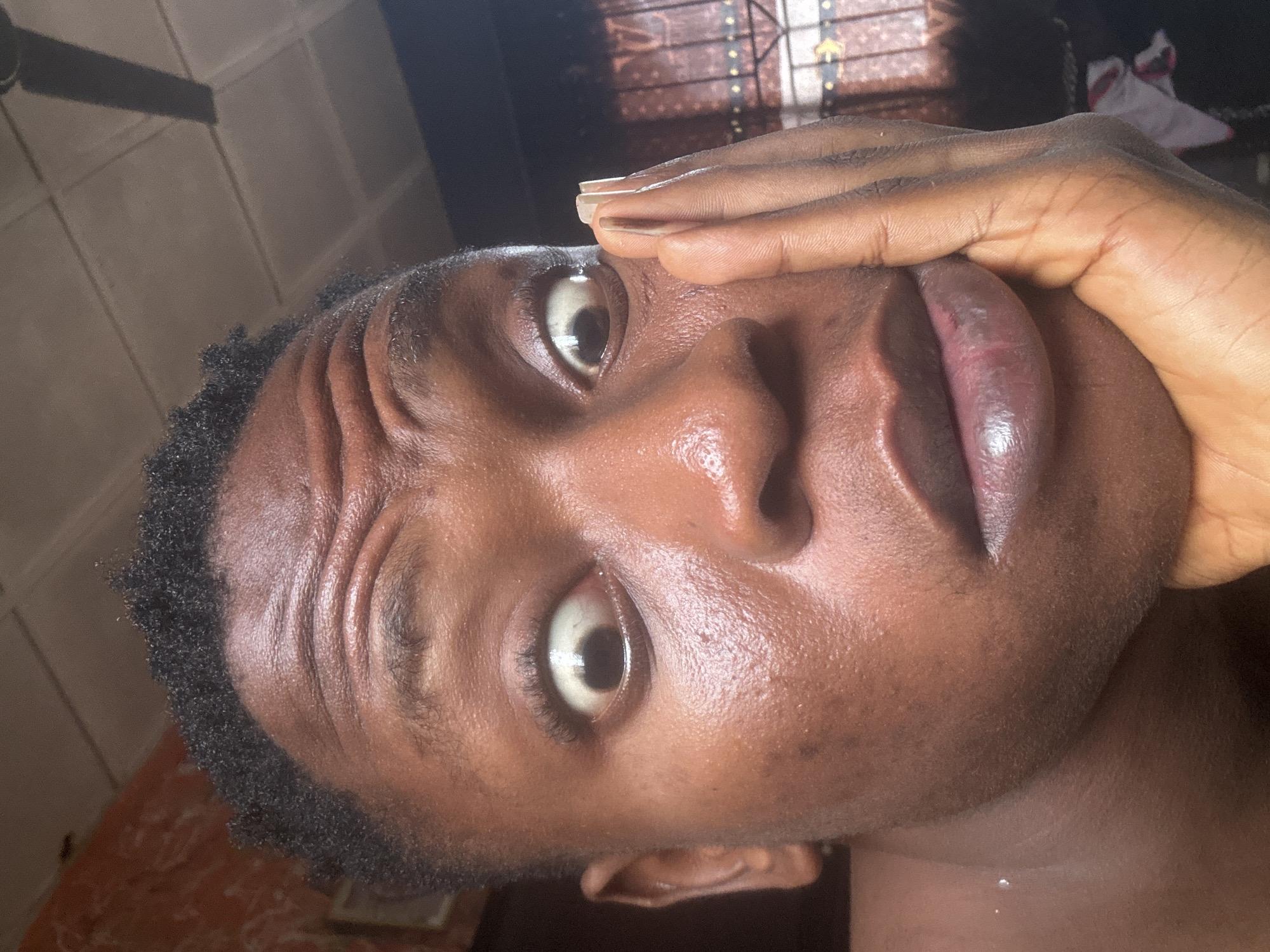 This is a close-up, eye-level selfie taken indoors, likely in a residential setting in Ikorodu, Nigeria. The subject, a dark-skinned individual with short, textured hair, is positioned slightly off-center to the right. Their face is the primary focus, with their left hand supporting their cheek. The subject's eyes are wide open and appear startled or surprised, with prominent wrinkles on their forehead suggesting a raised eyebrow expression. Their lips are slightly parted.
The background is dimly lit and shows signs of domestic life. To the upper left, a patterned wall with a framed picture is visible. Above the subject's head, a ceiling fan is silhouetted against a tiled ceiling. To the right, a window is covered by a patterned curtain with horizontal bars. The lighting appears to be natural, coming from the right, casting shadows and highlighting the sheen of the subject's skin, possibly indicating sweat or a product applied to the face. The overall impression is one of an impromptu captured moment of surprise or a strong emotional reaction.