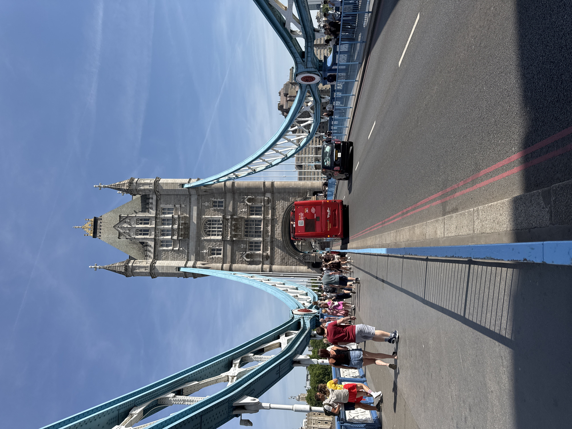 The image shows the Tower Bridge in London, United Kingdom. The view is from the bridge itself, looking towards one of the bridge's towers. There is a red double-decker bus and a black car on the roadway. Pedestrians are walking along the side of the bridge. The sky is blue with some light clouds. The bridge structure is painted in blue and white.