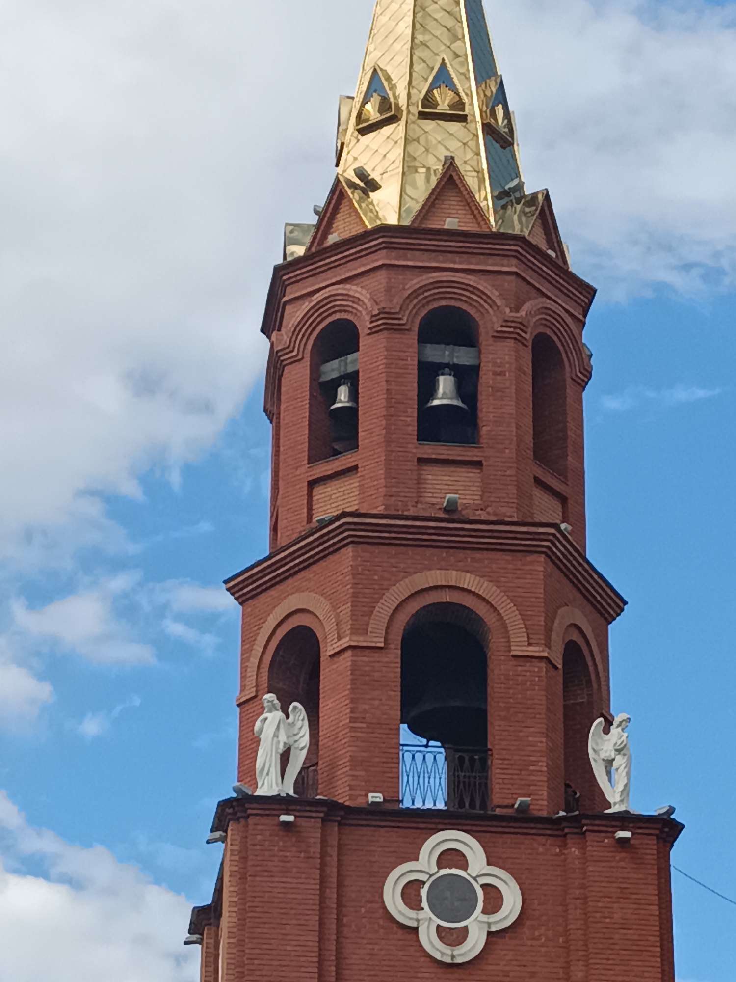 The image depicts the top of a brick bell tower with a gold-colored spire, set against a backdrop of a blue sky with white clouds. The tower has arched openings containing bells. There are white statues of angels on the tower's upper section. A decorative element, resembling a four-leaf clover with a central circle, is visible on the brick facade. The tower is the bell tower of St. George Church in Chaikovsky, Russia.