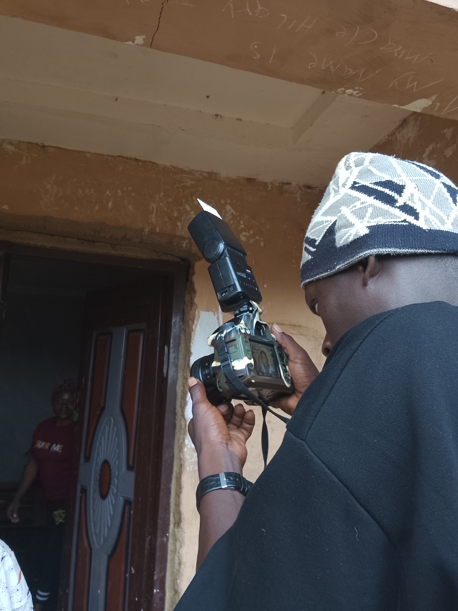 The photograph depicts a person wearing a patterned skullcap and a black shirt or jacket, holding a digital SLR camera with a flash attachment. The person is reviewing a picture on the camera screen. In the background, there is a doorway and a woman standing in the doorway wearing a red shirt. The walls surrounding the doorway appear to be painted in a tan or beige color. There are faded inscriptions in white on the wall above the doorway. The image appears to have been taken outdoors in the city of Unknown, Unknown.