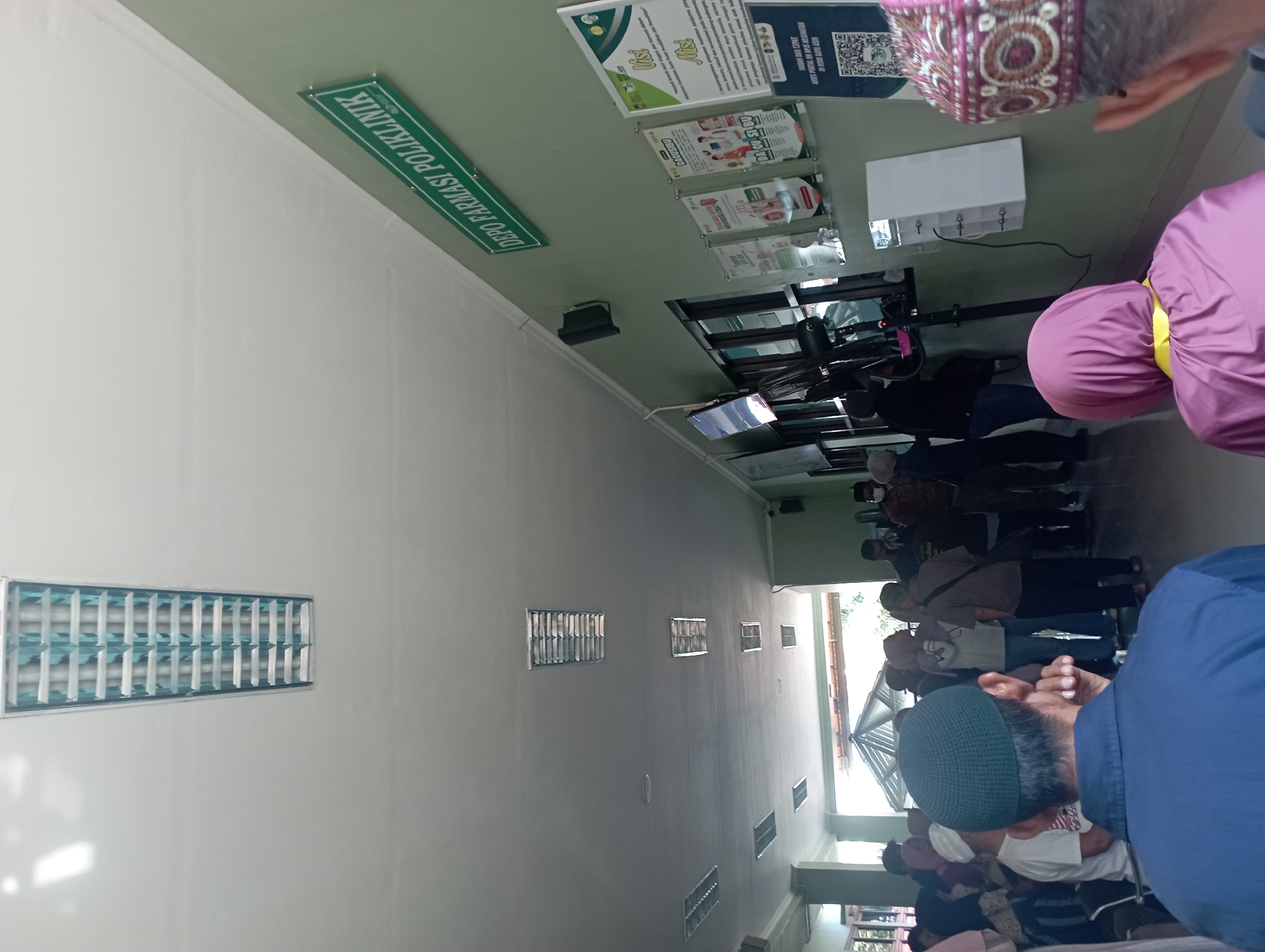 The image captures a scene inside what appears to be a public facility, possibly a health clinic or government office, located in Purwakarta, Indonesia. In the foreground, a person wearing a blue shirt and a dark green skullcap is seen from behind, facing away from the camera. To their right, another individual, partially visible, wears a maroon and white patterned peci hat. In the middle ground, a crowd of people are gathered, some standing and some possibly seated, indicating a queue or waiting area. They are diverse in dress, with many individuals wearing headscarves. The environment features a long, light-colored hallway or waiting room. The walls are painted a pale green. A prominent green sign with white text reads "DEPO FARMASI POLIKLINIK," suggesting the presence of a pharmacy within a polyclinic. Several framed posters with text and graphics are displayed on the wall, likely containing information or advertisements. Electrical outlets and what appear to be screens or monitors are also visible along the wall. Fluorescent light fixtures are embedded in the ceiling, illuminating the space. The overall impression is a busy, functional indoor space designed to serve a number of people.
