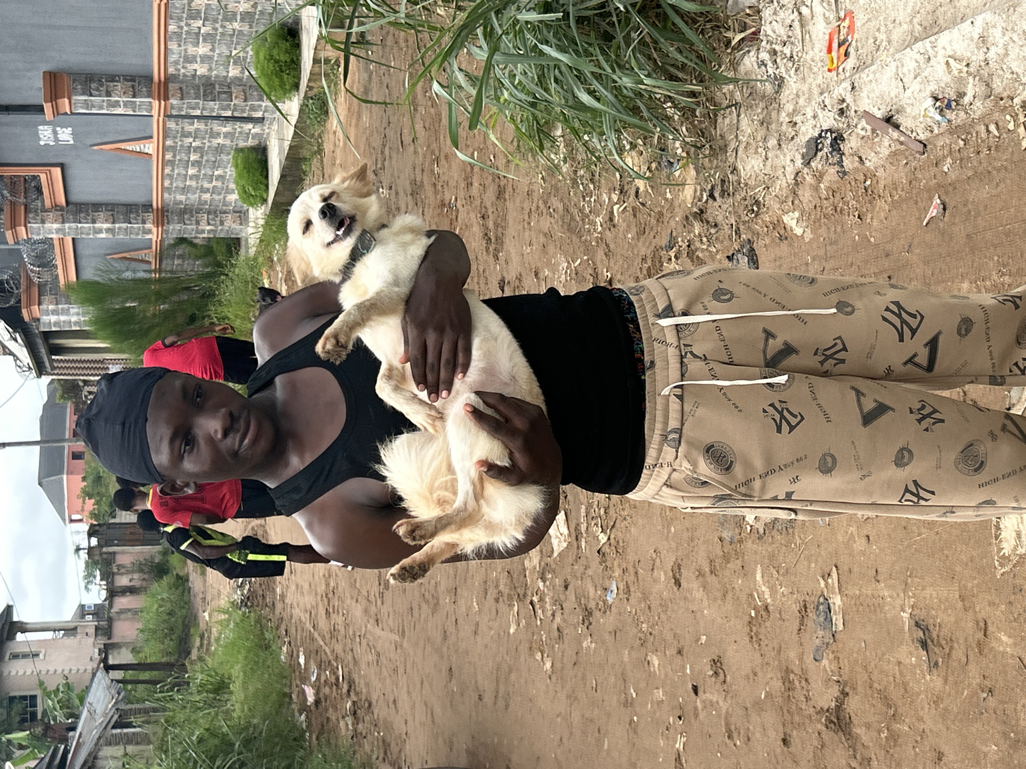 A young man wearing a black durag and a black tank top is holding a light-colored dog upside down in his arms. He is wearing tan sweatpants with a repeating pattern of logos and text. The dog appears to be happy and is panting with its tongue out. In the background, there are other people walking on a dirt road, some buildings, and dense green vegetation. The location is identified as Ihiagwa, Nigeria. The environment appears to be an outdoor urban or semi-urban setting with a dirt road and some scattered debris.