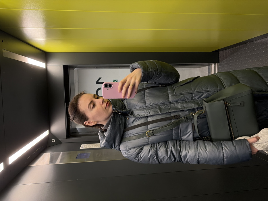 The scene depicts an interior view of an elevator in Ковалёво, Russia. A female subject is shown taking a mirror selfie, holding a pink-cased smartphone in her right hand, which displays a ring on the fourth finger. Her left hand grasps a folded light-colored garment. She wears a long, dark grey puffer coat over a grey turtleneck, and her dark brown hair is partially tied back. A dark green shoulder bag with a visible front zipper pocket is strapped over her left shoulder. The elevator features dark grey vertical paneling on the left and rear walls, with bright yellow vertical panels on the right. Overhead, two linear LED light fixtures illuminate the space. A small, circular security camera is mounted on the rear wall, above the dark elevator door frame. Through the partially open elevator door in the background reflection, a lighter-colored hallway is visible, featuring a large dark graphic symbol and a green illuminated exit sign on a white wall. A control panel with illuminated blue buttons is partially visible on the left wall.